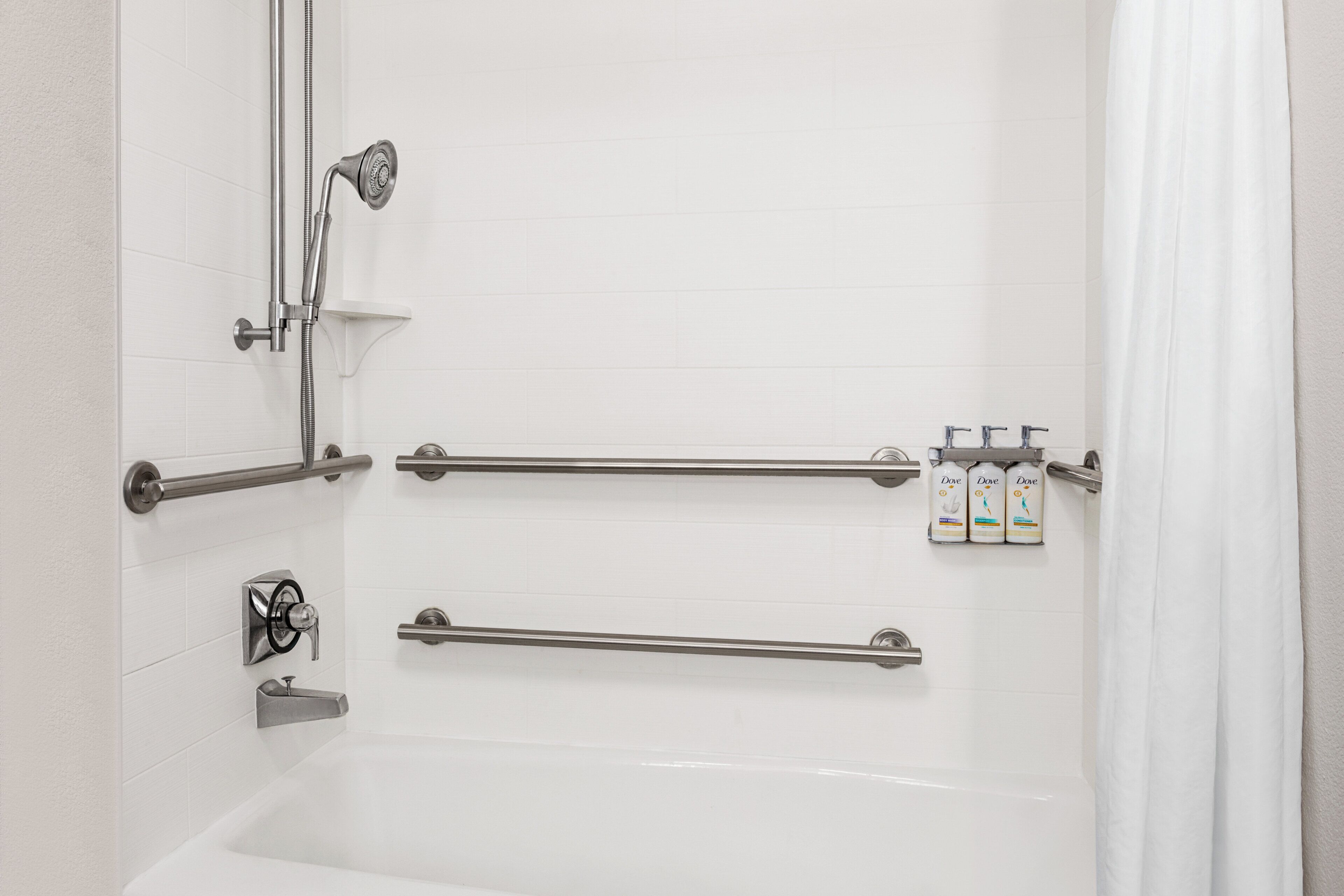 A bathroom with a bathtub, grab bars, a showerhead, and toiletries.