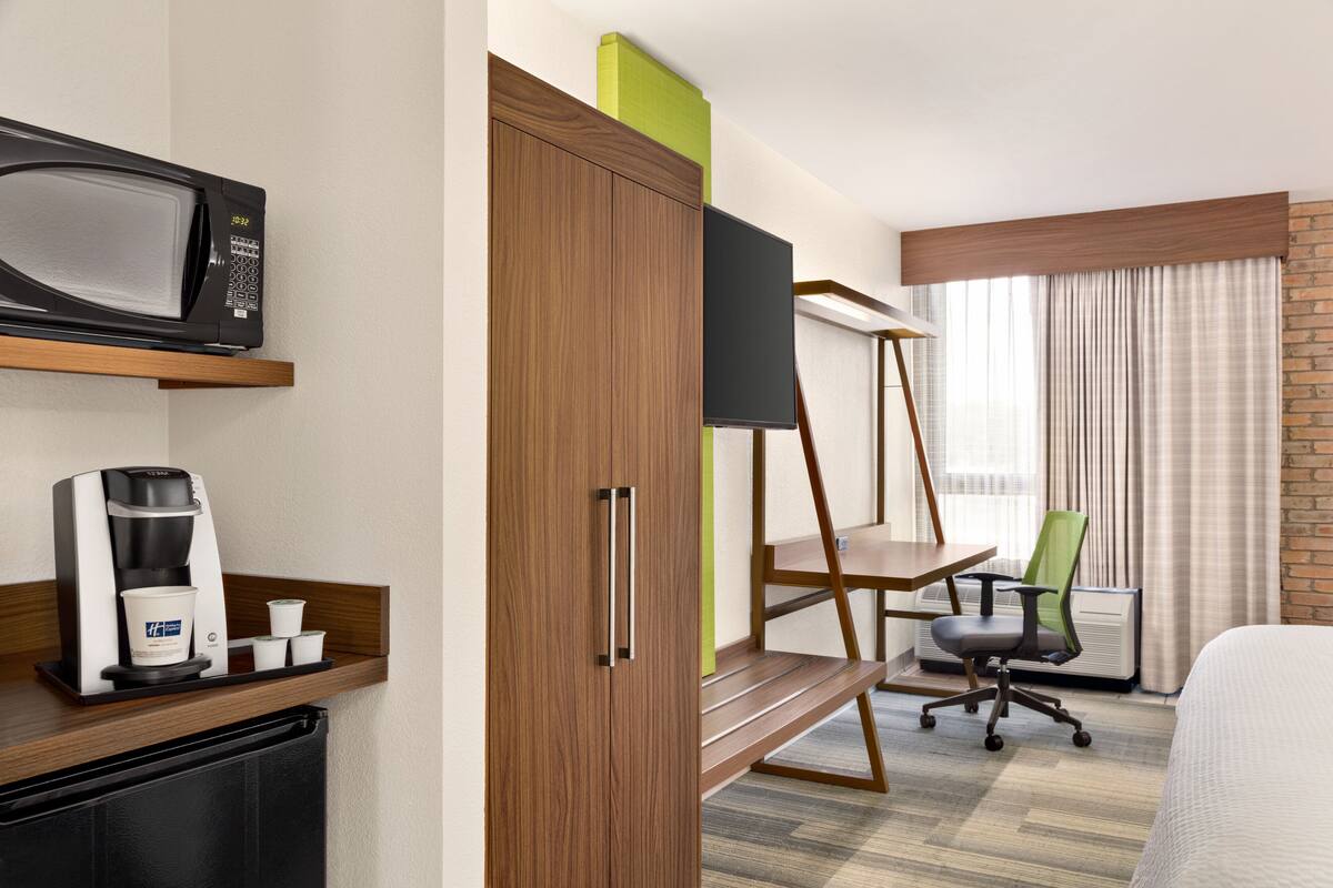 Habitación de hotel con cama, escritorio con silla, microondas, cafetera y televisor sobre un mueble.
