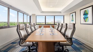 Un salón de conferencias con una larga mesa, varias sillas y grandes ventanales que ofrecen vistas a zonas verdes y edificios.