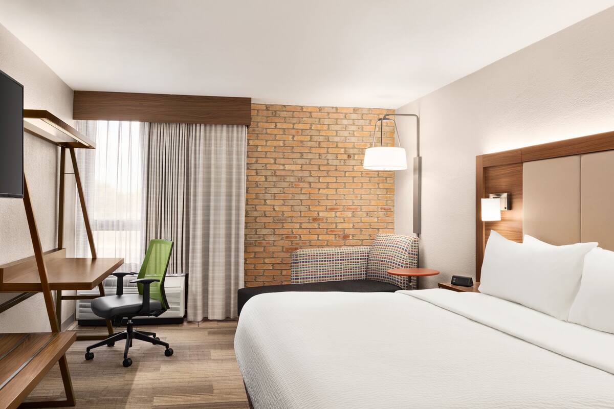Habitación de hotel con cama, escritorio, silla, televisor, lámpara y pared de ladrillo.