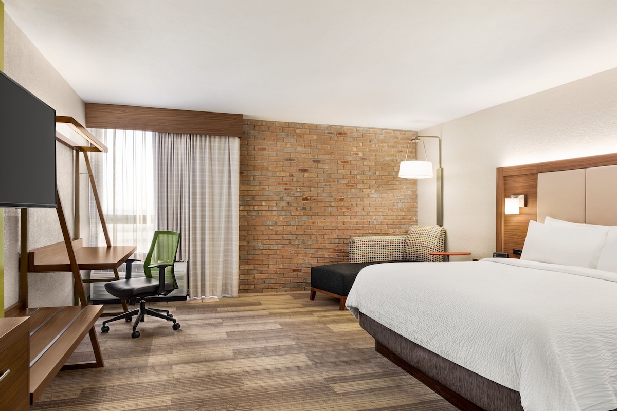 A hotel room with a brick accent wall, a bed with white linens, a desk with a chair, and a television.