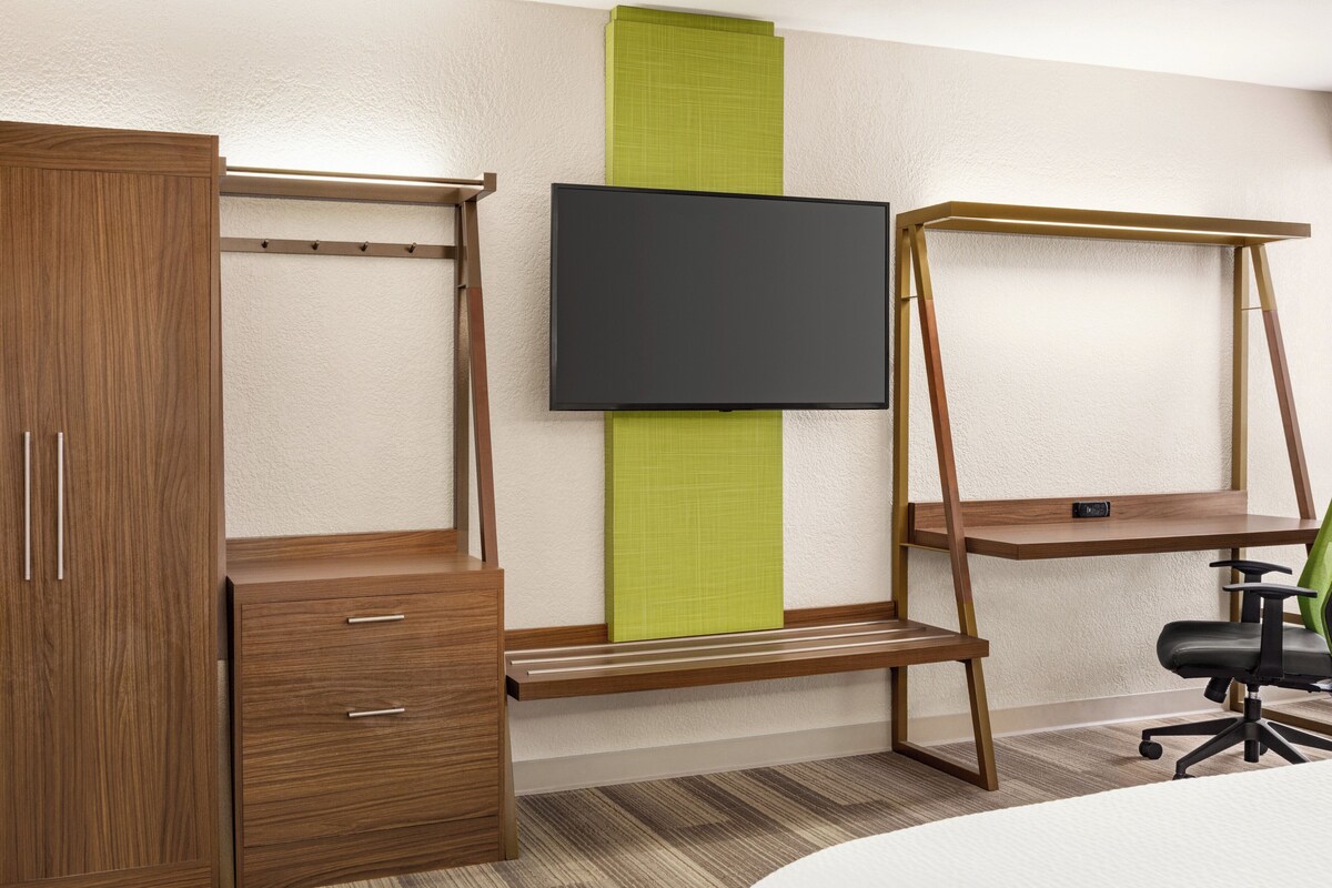 Ein Zimmer mit einem Holzschrank, einem Schreibtisch mit einem vertikalen grünen Akzent, einem schwarzen Stuhl und einem fest montierten Flachbildfernseher.