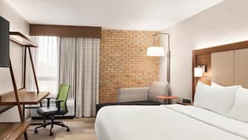 A hotel room with a bed, a desk, a chair, a TV, a lamp, and a brick wall.