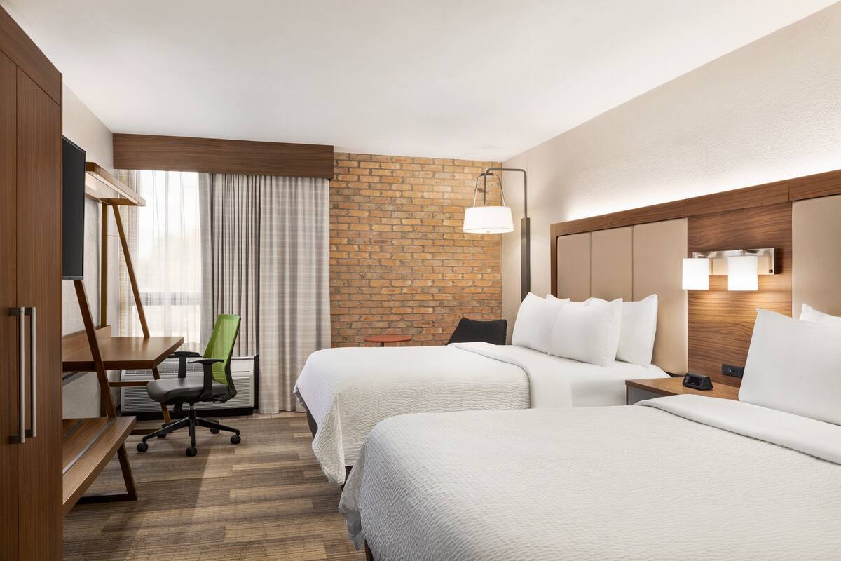 Habitación de hotel con dos camas, un escritorio, una silla y una pared de ladrillo decorativa.