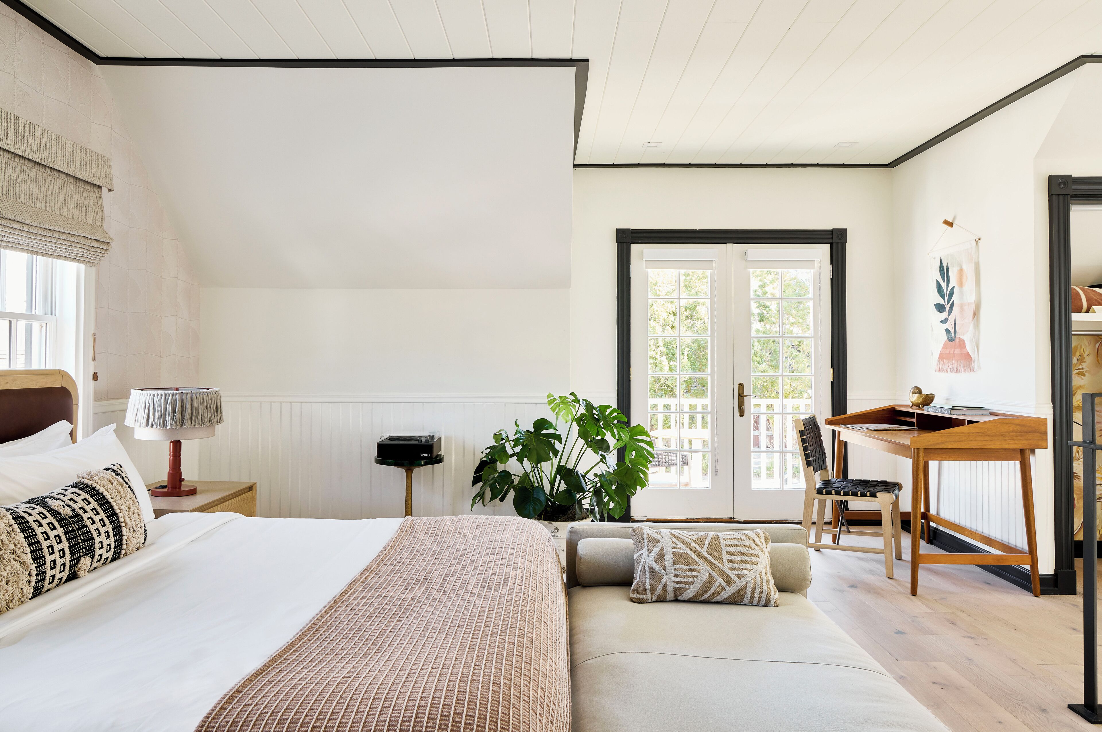 A bedroom with a bed, bedside table, lamp, potted plant, and a desk with a chair.