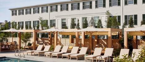 A poolside area with lounge chairs and umbrellas, a pool, and a building with multiple windows and a flat roof.