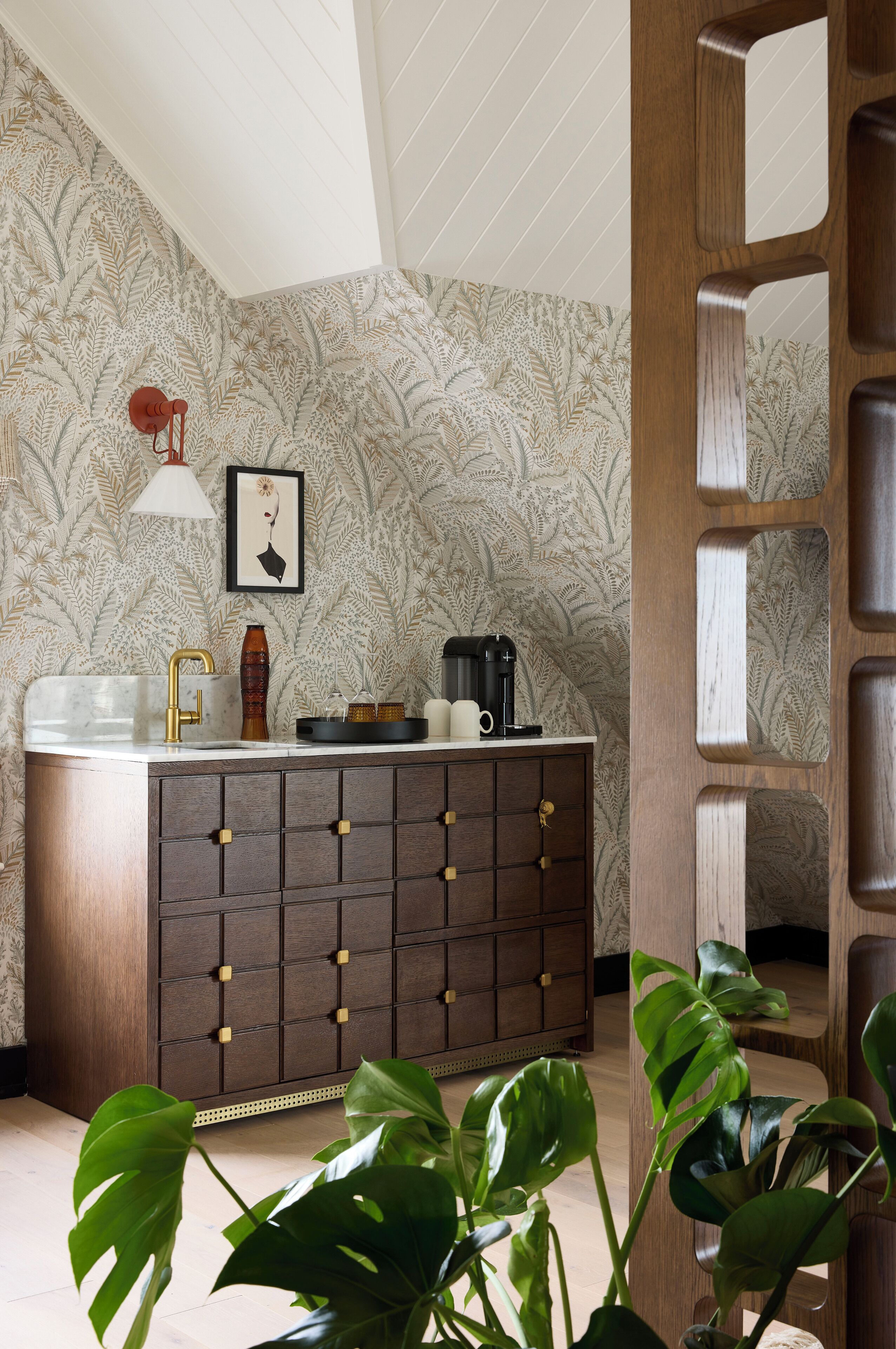 A bathroom with a patterned wallpaper, a wooden cabinet with drawers, a sink with a gold faucet, a coffee maker, and a framed picture on the wall.