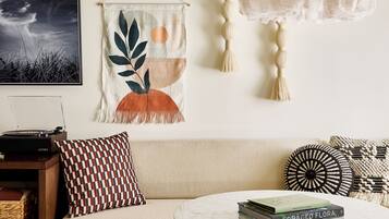 A living room with a sofa, a round coffee table, a wall-mounted macrame piece, a wall clock, and a record player.