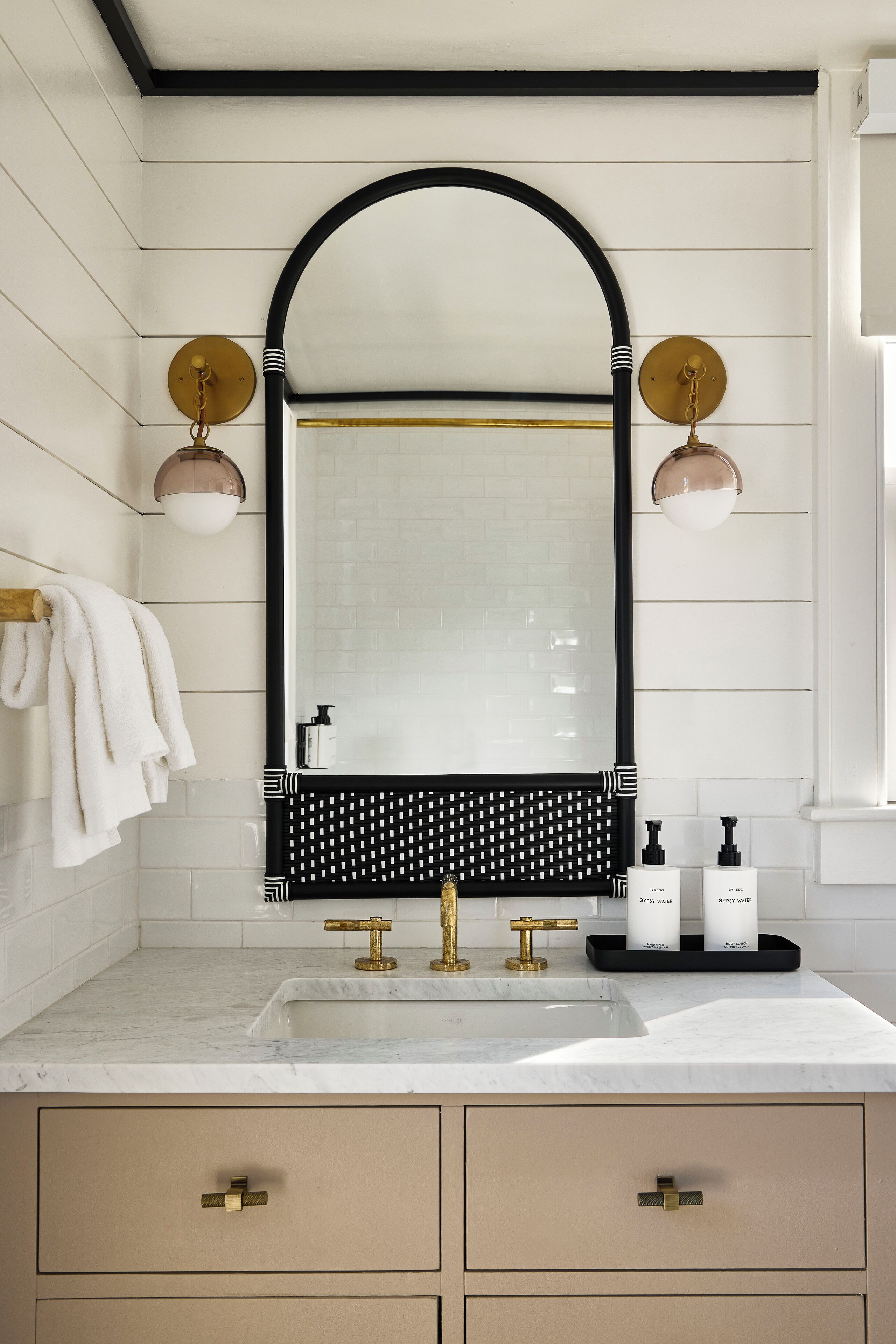 A bathroom with a white tiled wall, a black-framed mirror, a gold faucet, and two wall-mounted light fixtures.