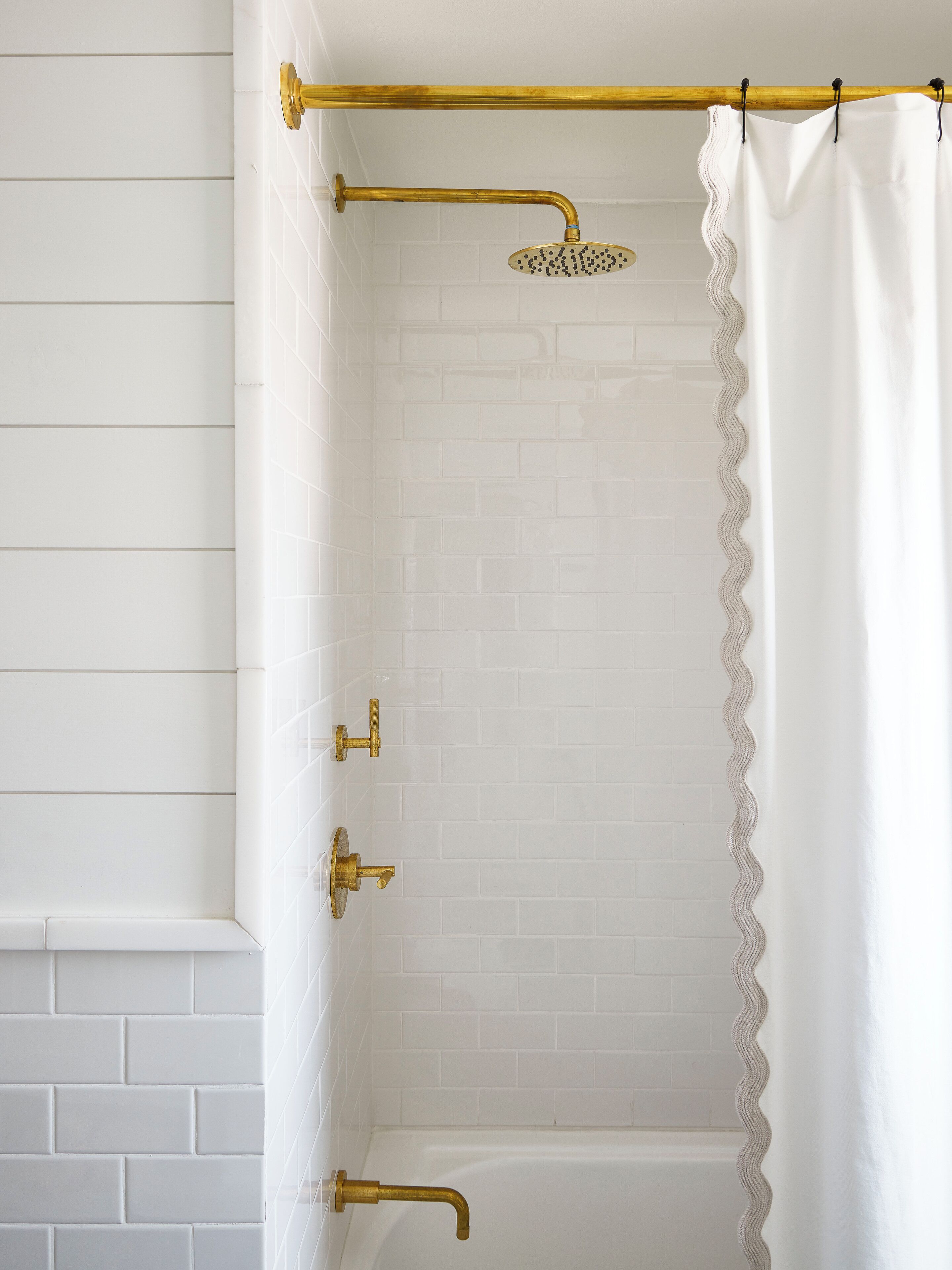 Un espace douche avec des murs recouverts de carreaux blancs, une pomme de douche en laiton et un rideau de douche blanc à bords ondulés.