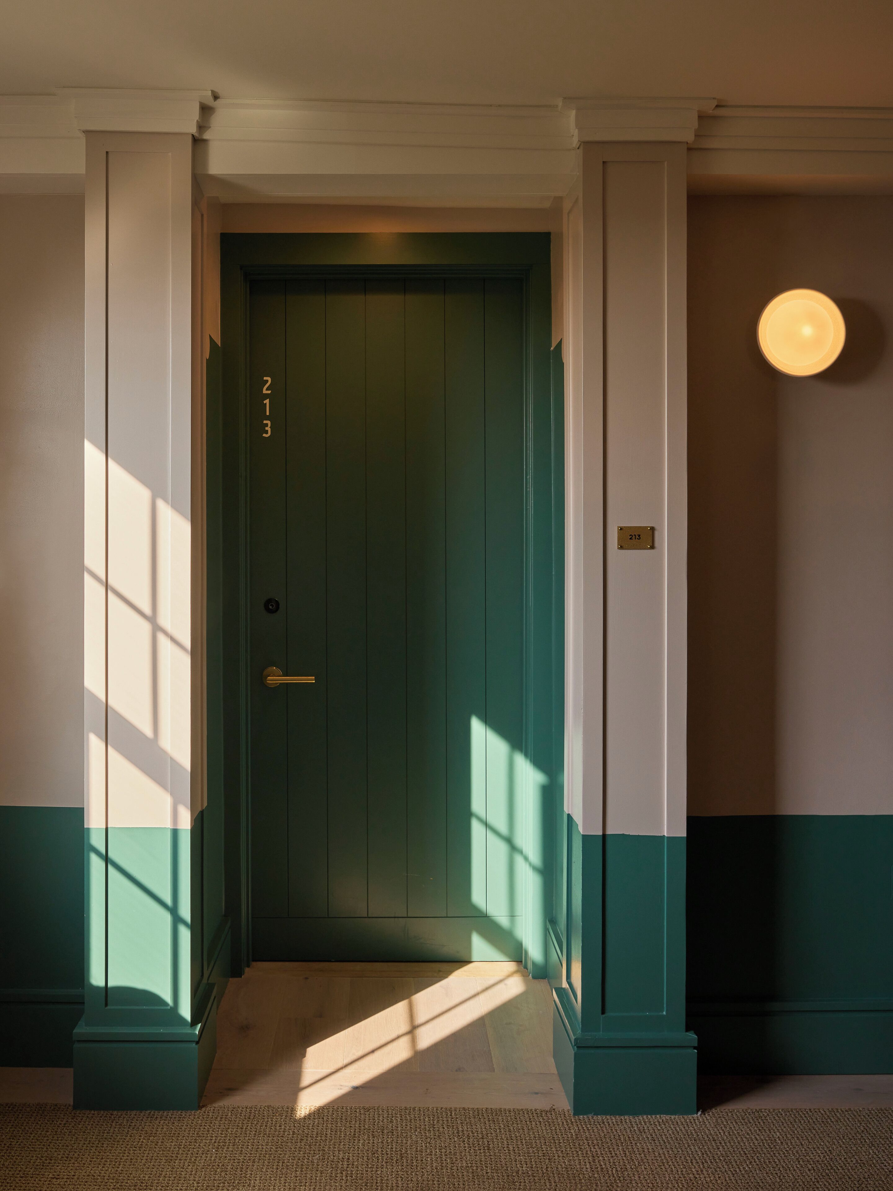 Una puerta verde con manija de latón, una placa con número y un accesorio de luz en la pared.