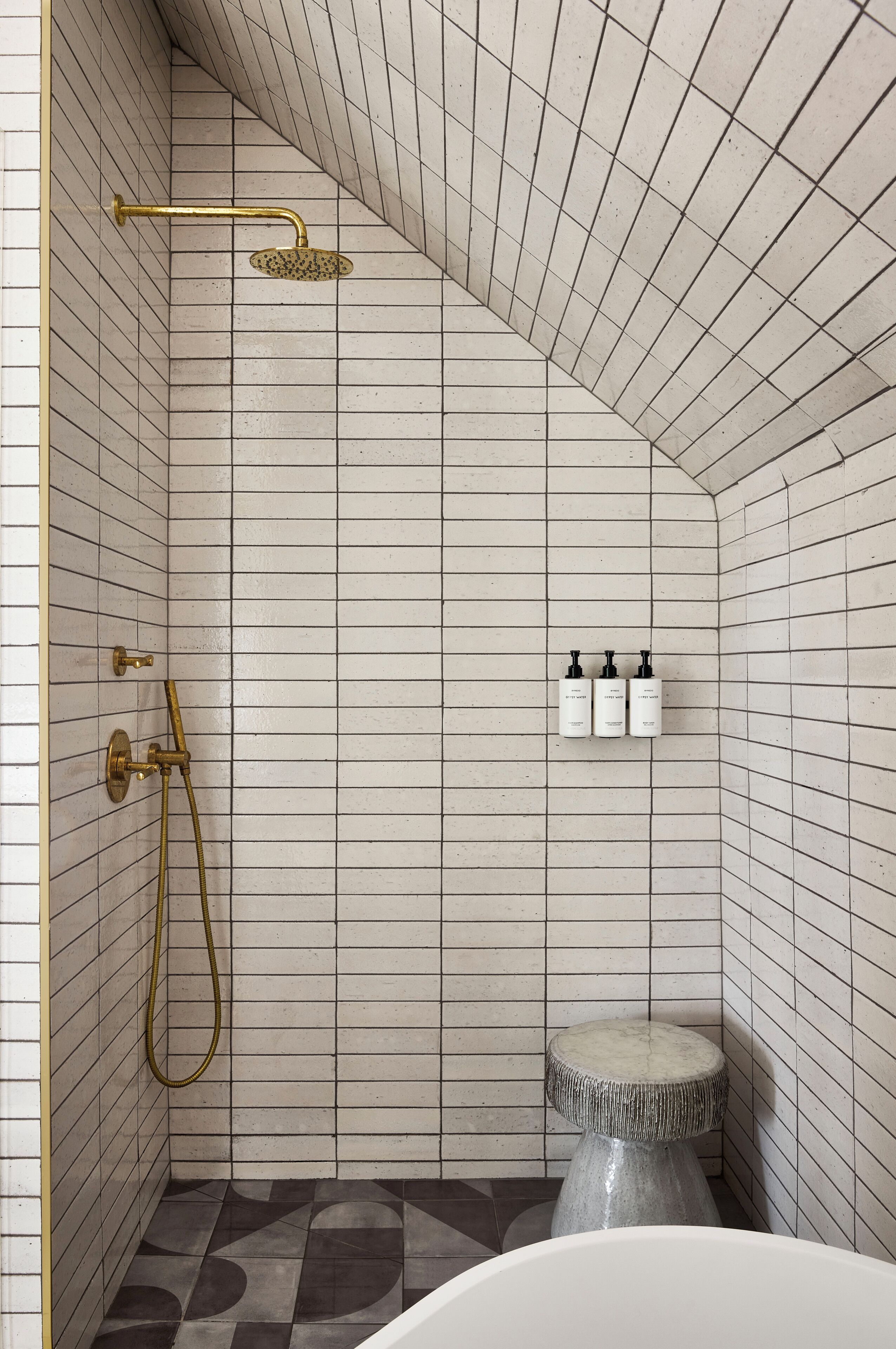 A bathroom with white tiled walls, a shower with a gold fixture, and a small round stool.