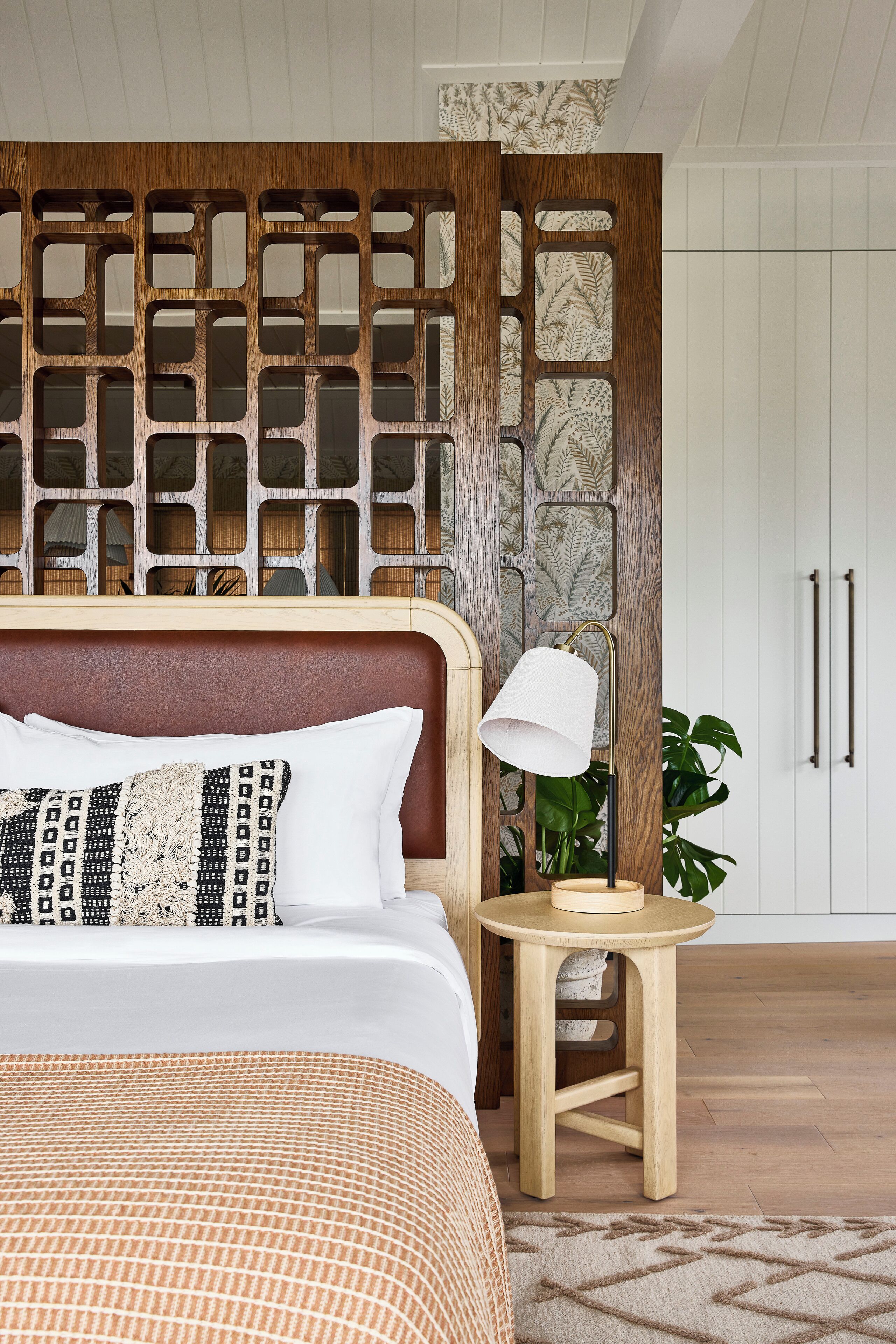 A neatly made bed with patterned pillows, a wooden headboard, a small wooden stool with a lamp, and a potted plant.