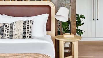 A neatly made bed with patterned pillows, a wooden headboard, a small wooden stool with a lamp, and a potted plant.