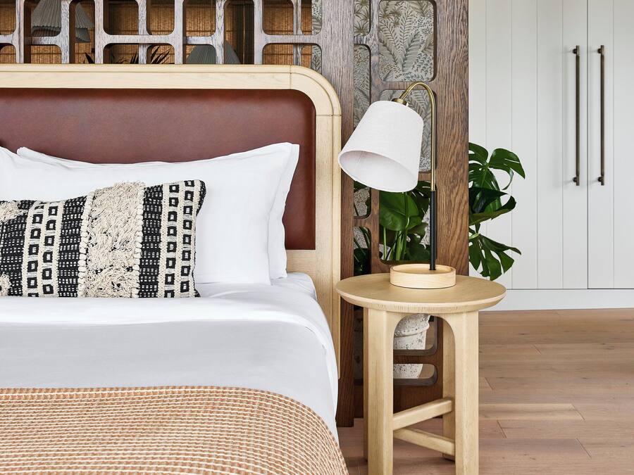 Ein ordentlich bezogenes Bett mit gemusterten Kissen, ein hölzernes Kopfende, ein kleiner Holzschemel mit einer Lampe und eine Topfpflanze.