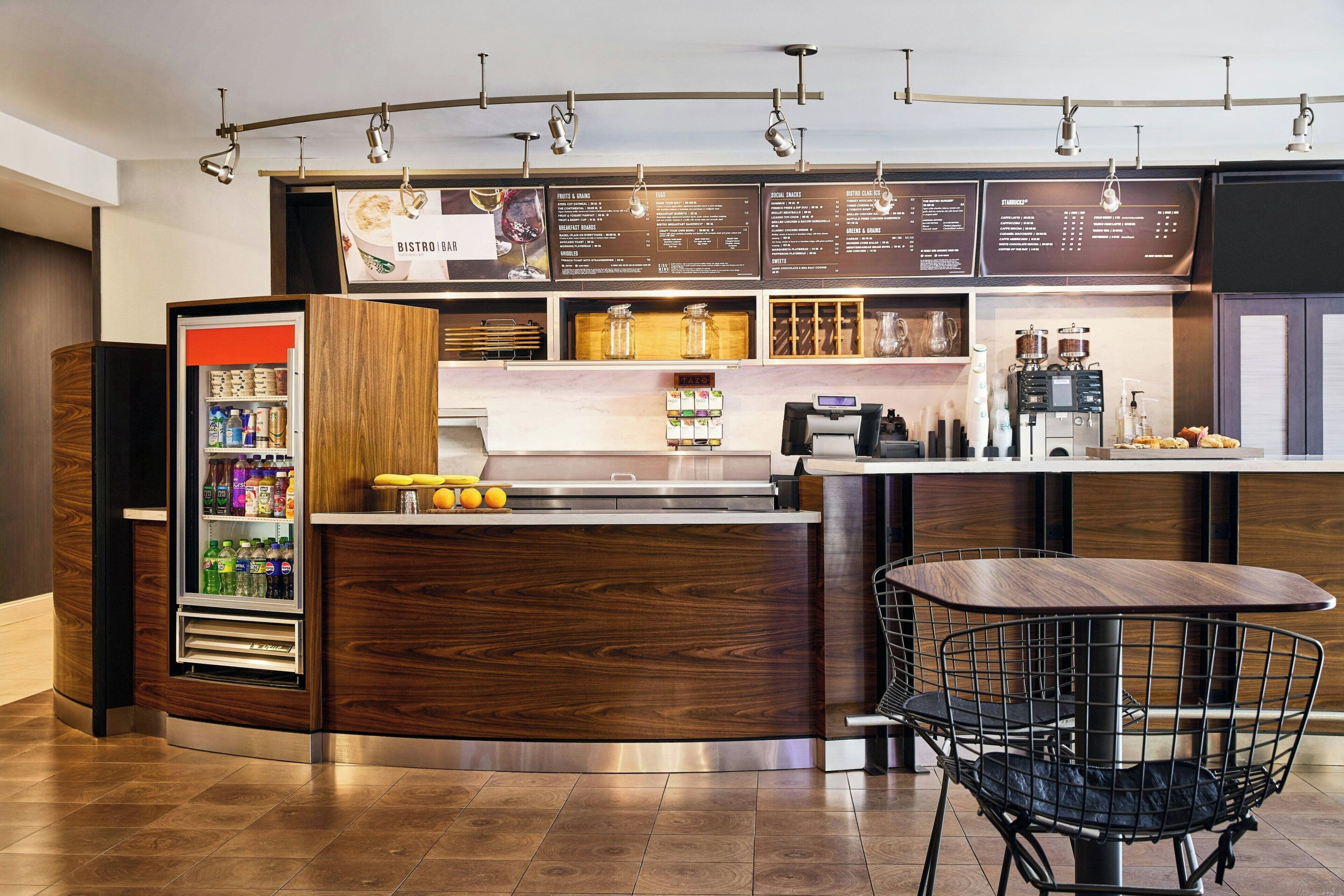 A modern bistro with a wooden counter, a refrigerated beverage dispenser, a coffee machine, and a metal chair.