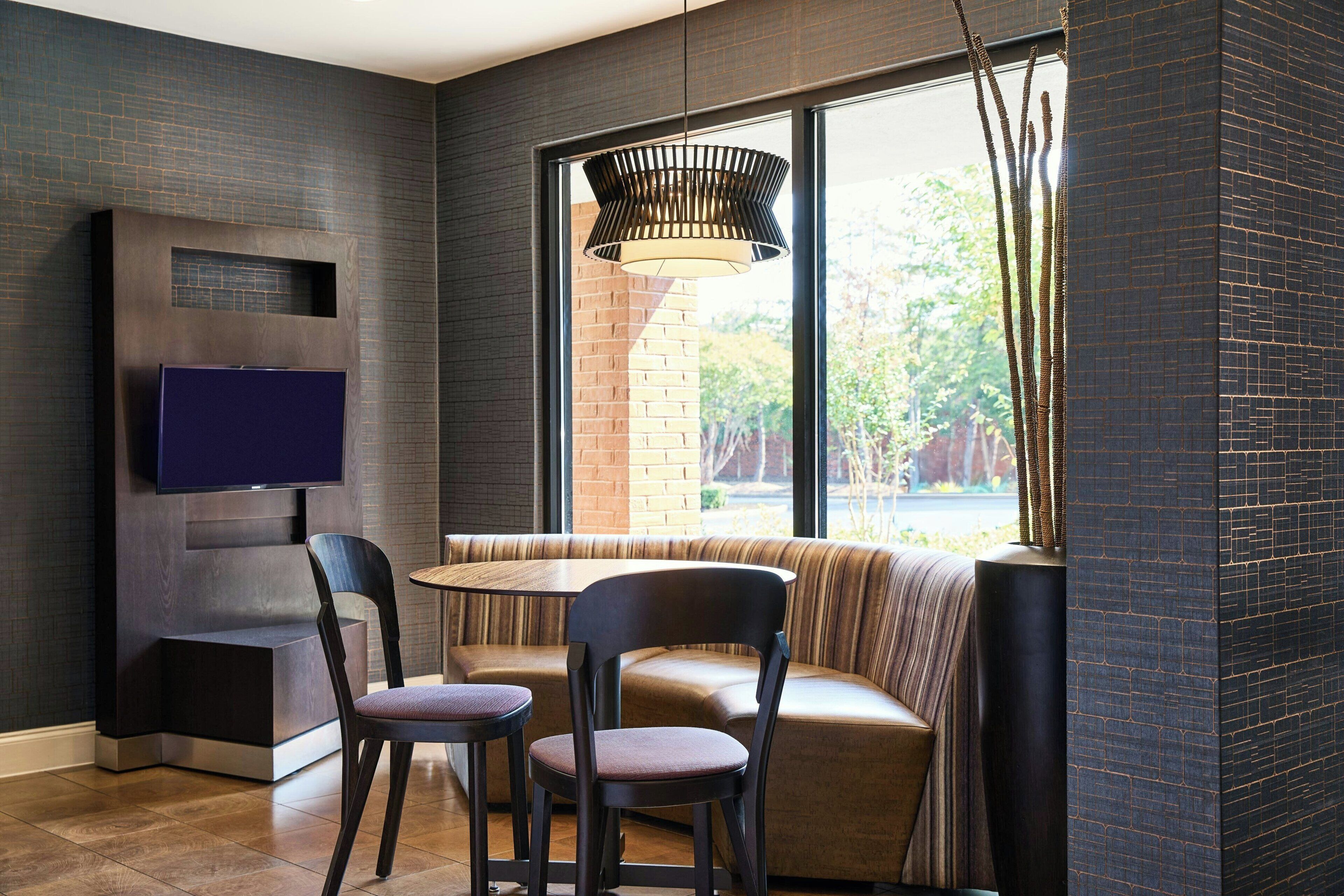 A modern interior with a TV mounted on a wall, a seating area with cushioned chairs, a round table, and a large window with a view of greenery.