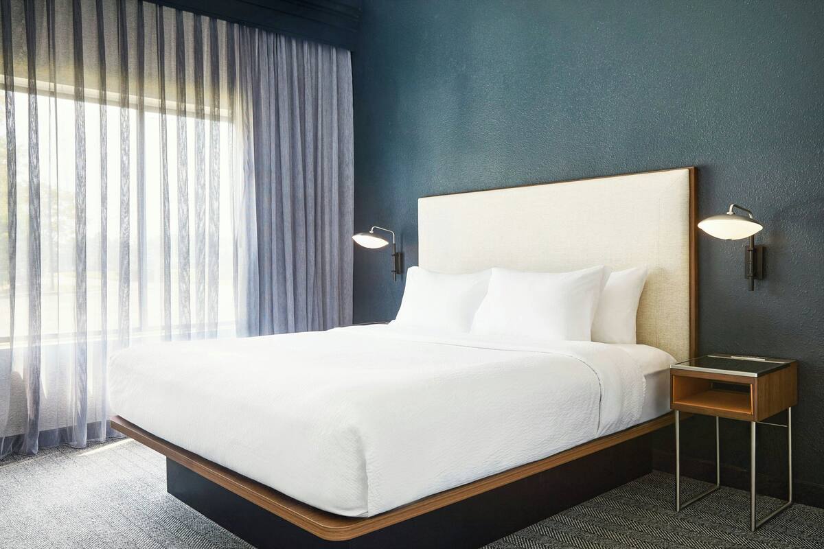A hotel room with a large bed, two bedside lamps, a nightstand, and a view of the outdoors through sheer curtains.