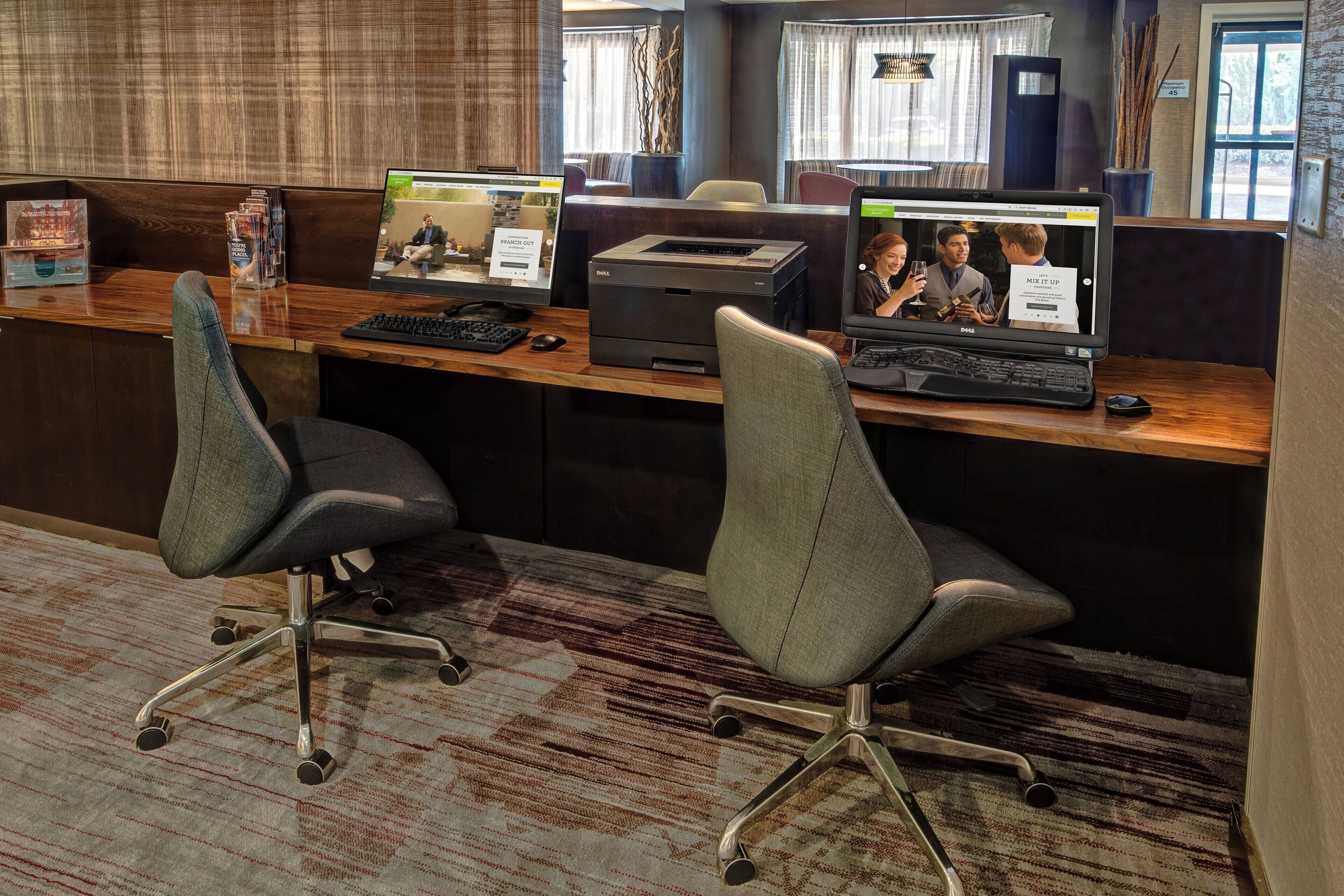 A reception desk with two office chairs, a desktop computer, a printer, and a large monitor displaying a video conference.