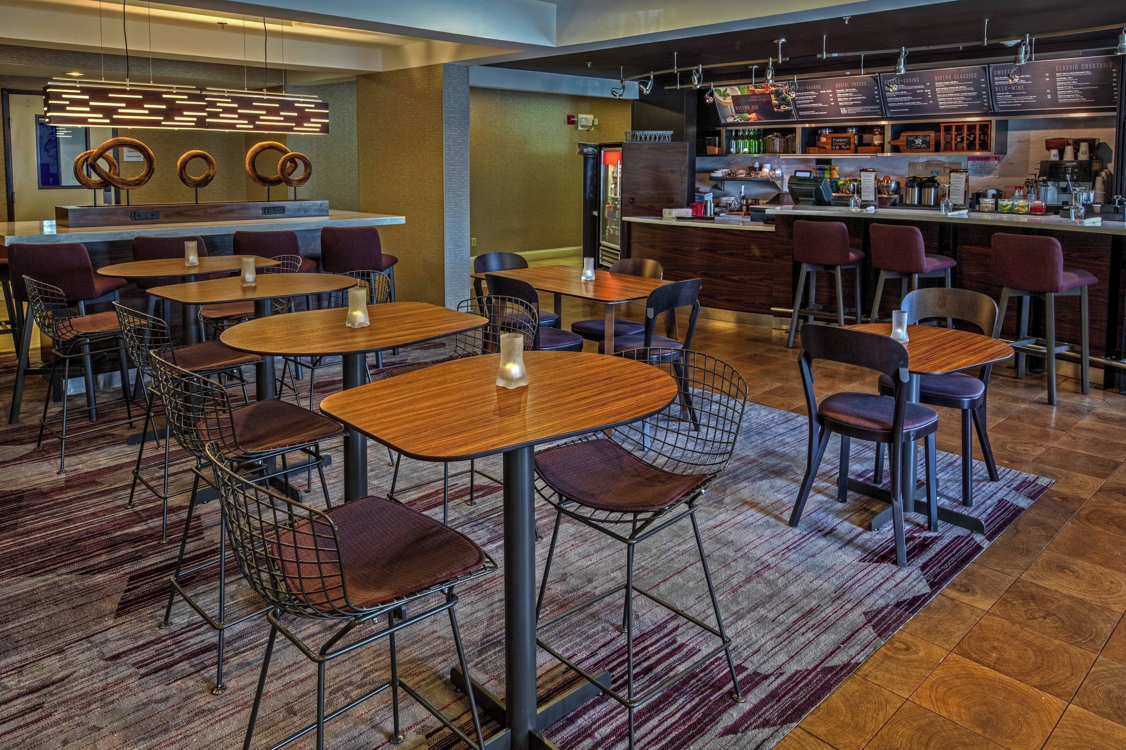 A modern restaurant with wooden tables and metal chairs, a bar area with a menu, and a patterned carpet.