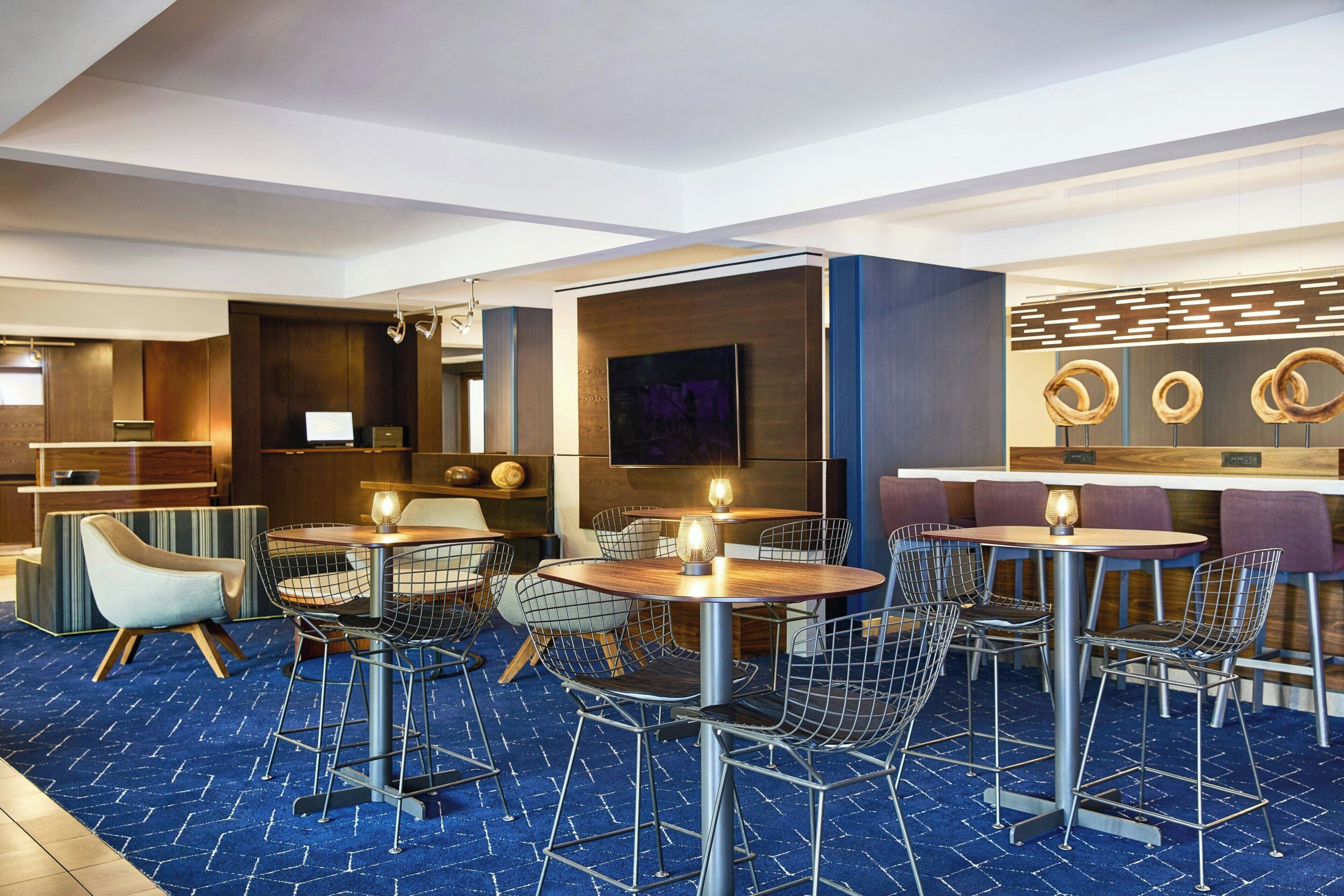 Un vestíbulo de hotel moderno con mesas redondas, sillas de metal, una zona de bar y una pantalla de televisión grande.