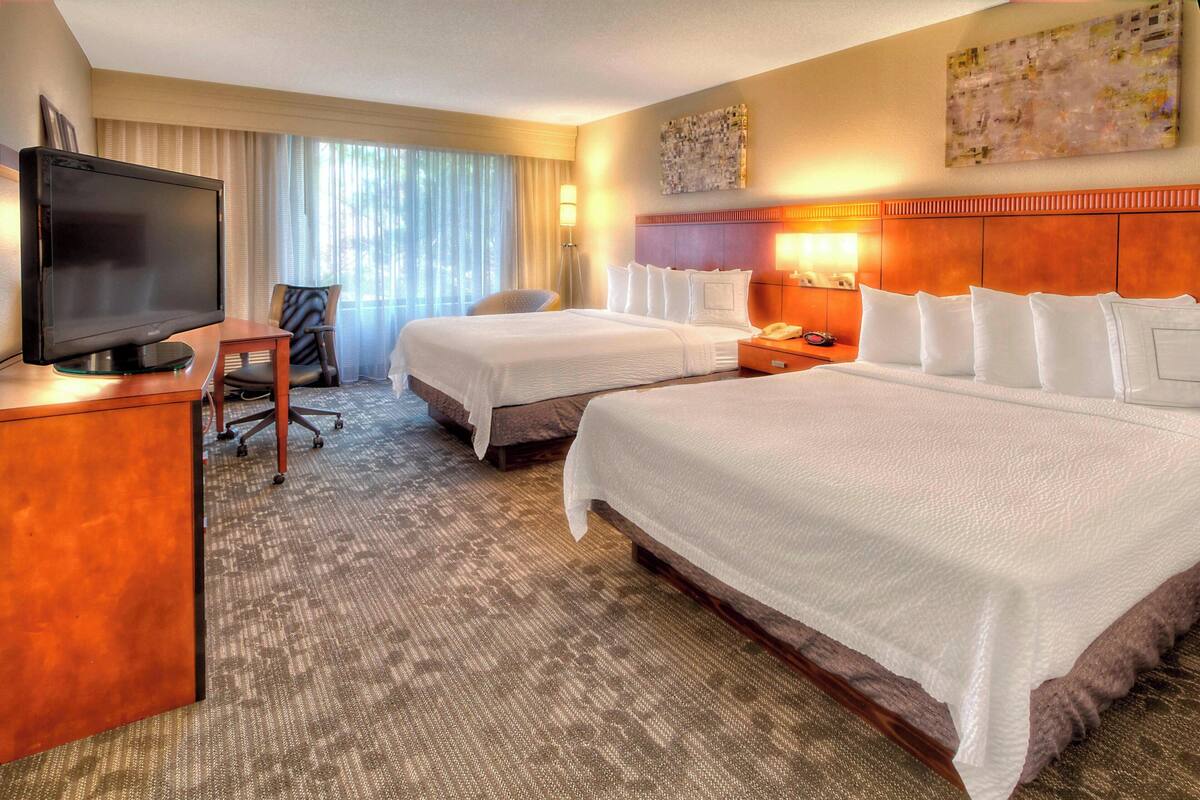 A hotel room with two beds, a desk with a television, a chair, and a large window with curtains.