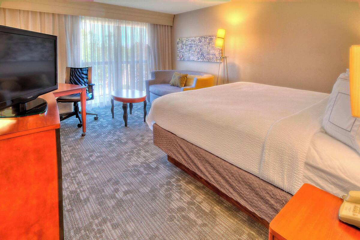 A hotel room with a bed, a desk with a television, a chair, a small table, and a lamp.