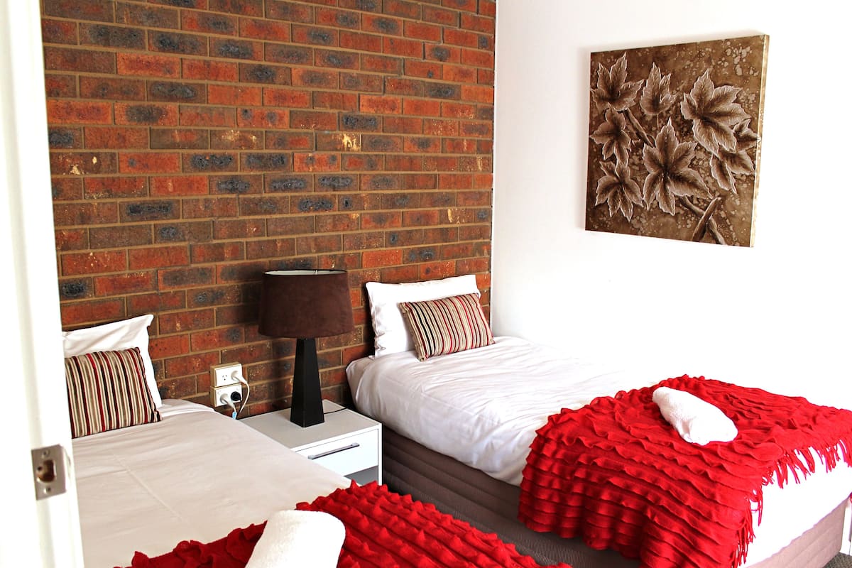 A hotel room with a brick wall, two beds with white bedding, red throw blankets, a nightstand with a lamp, and a painting on the wall.