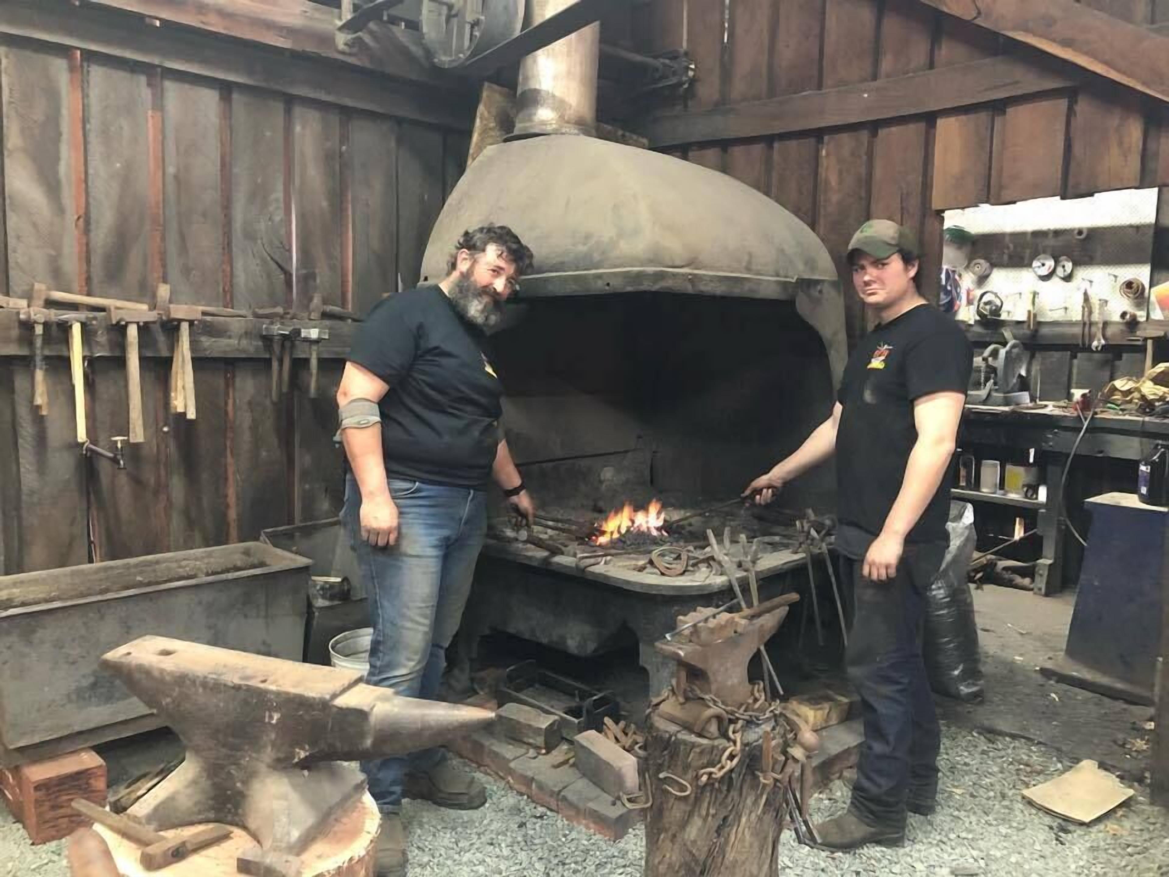 Two individuals working with a large, industrial forge in a workshop setting.