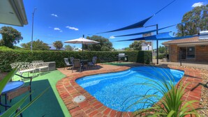 Una piscina con borde de ladrillo, rodeada por una terraza con mobiliario de exterior y un seto.
