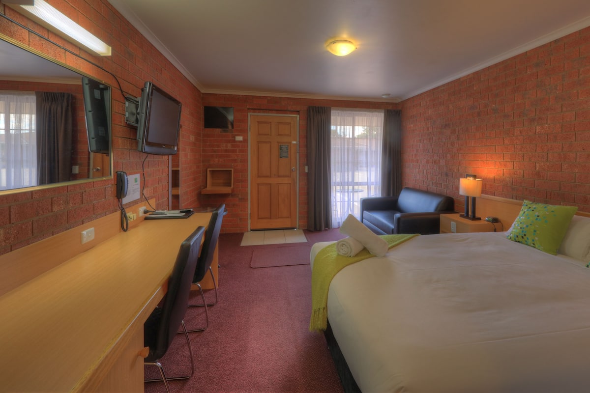 A hotel room with a bed, a desk, a chair, a TV, and a brick wall.