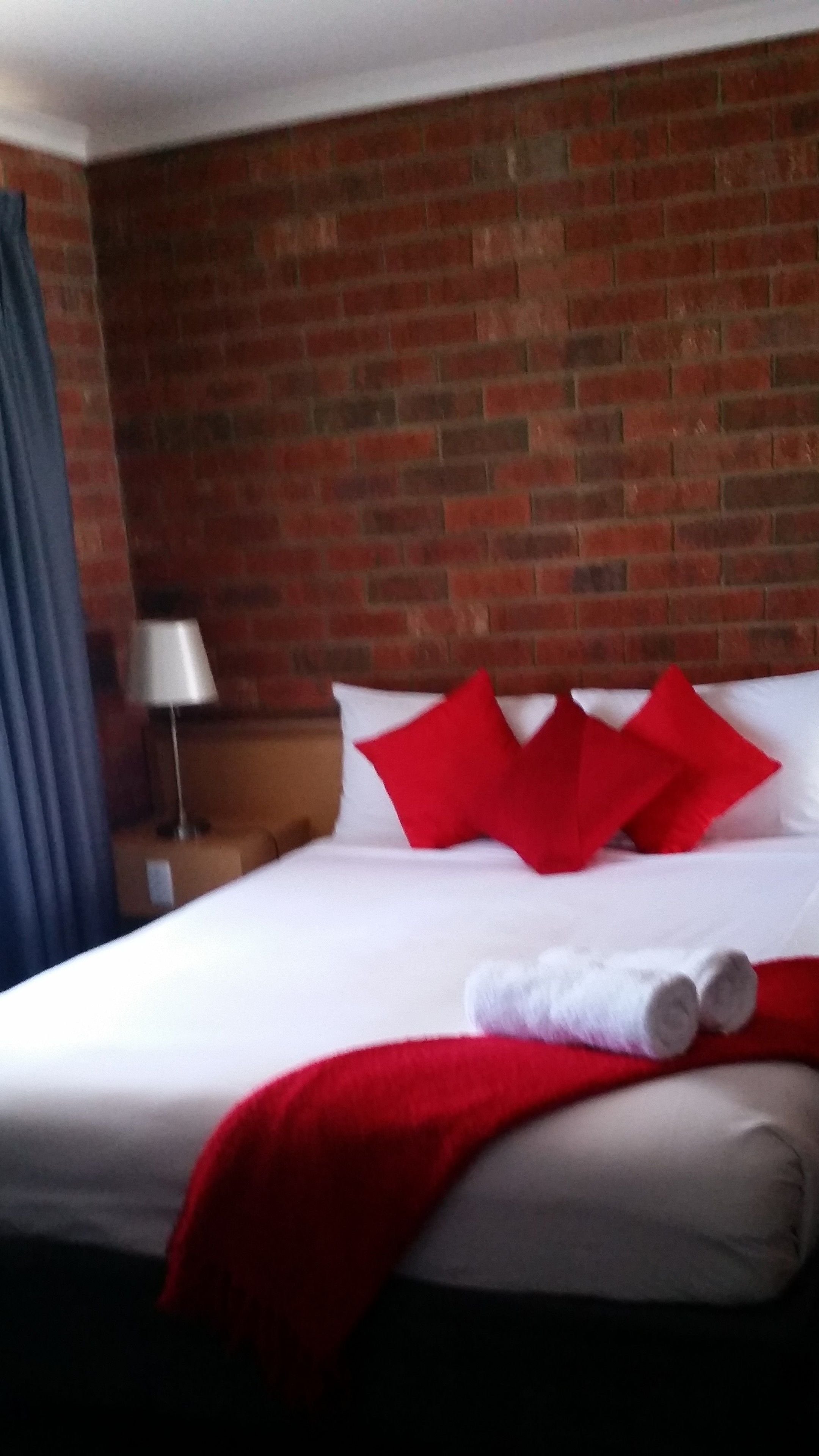 A hotel room with a brick wall, a bed with white bedding and red pillows, a nightstand with a lamp, and a red throw blanket.