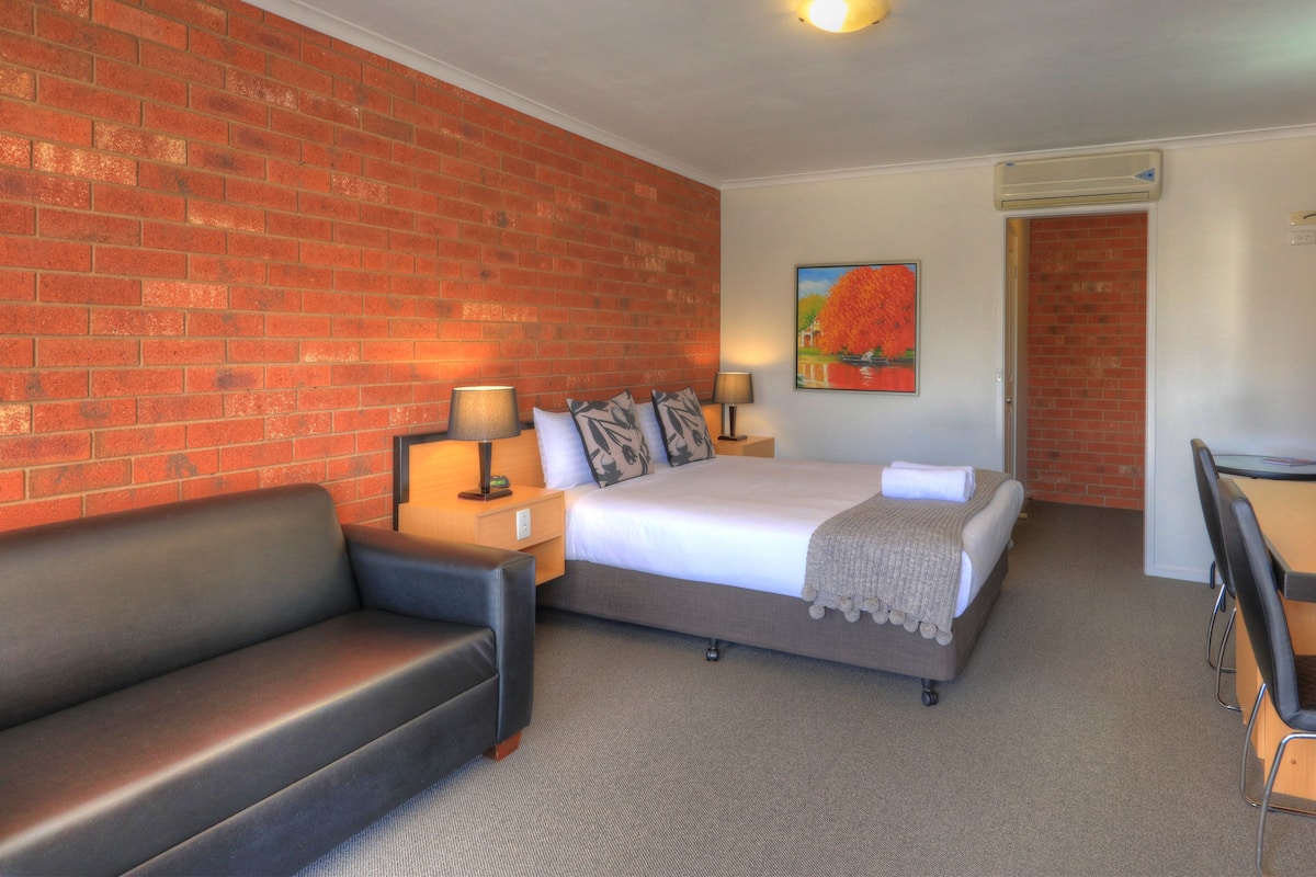 A hotel room with a brick wall, a bed with white bedding, a black leather sofa, a desk with a chair, and a painting on the wall.