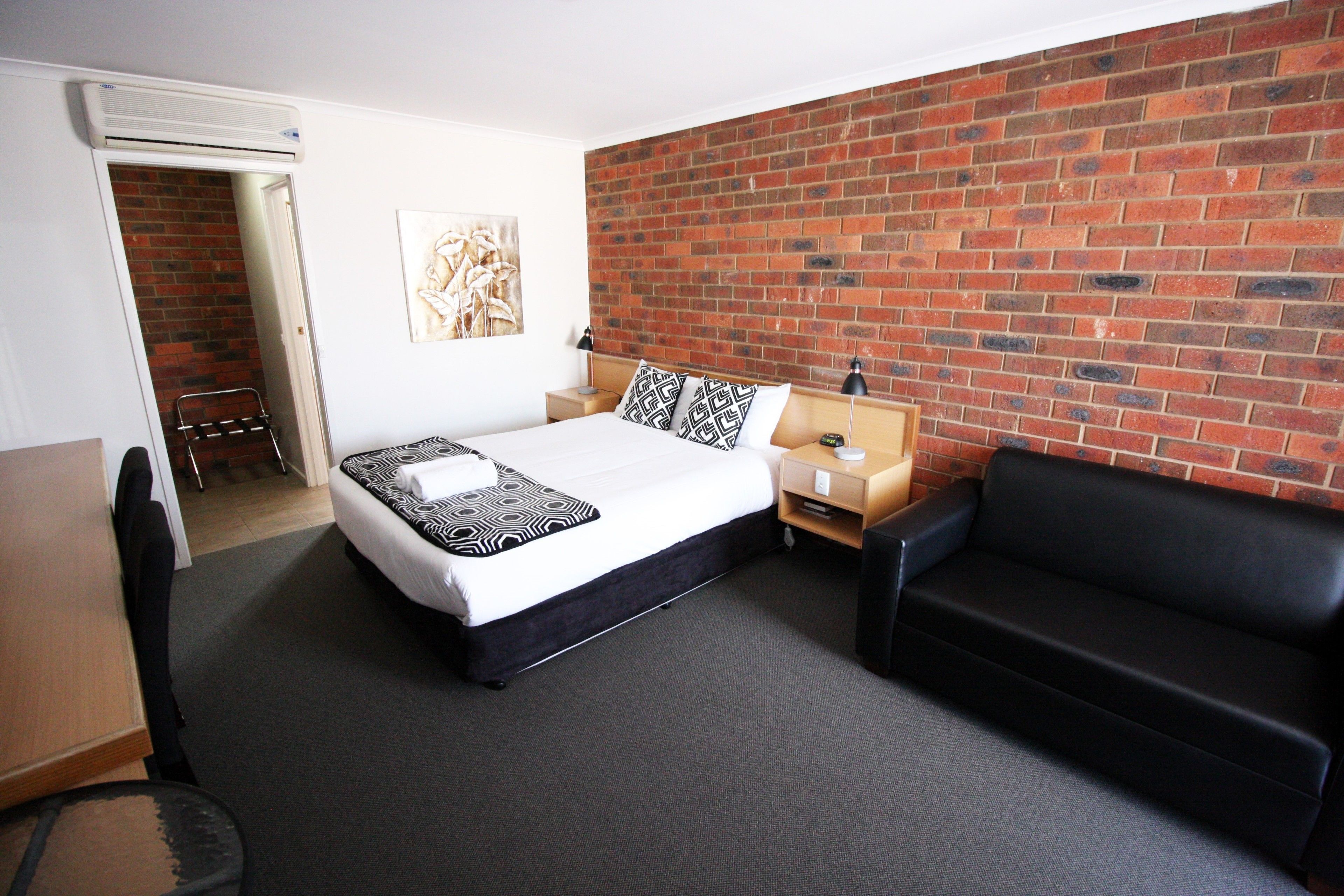 A hotel room with a bed, a black leather sofa, a brick wall, a small wooden nightstand, and a door leading to a bathroom.