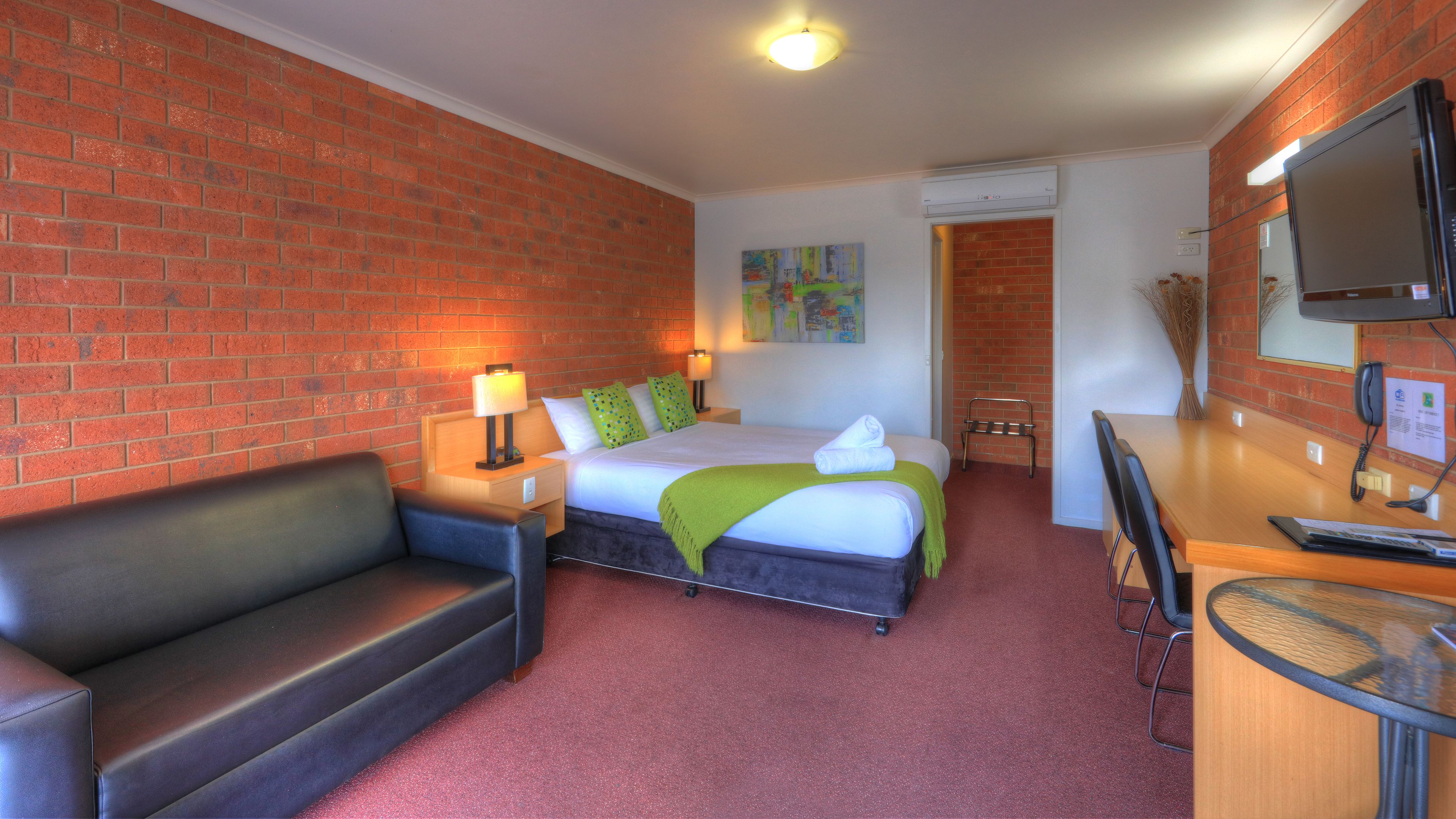 Habitación de hotel con pared de ladrillo, cama con ropa de cama verde, escritorio con teléfono y televisor en la pared.