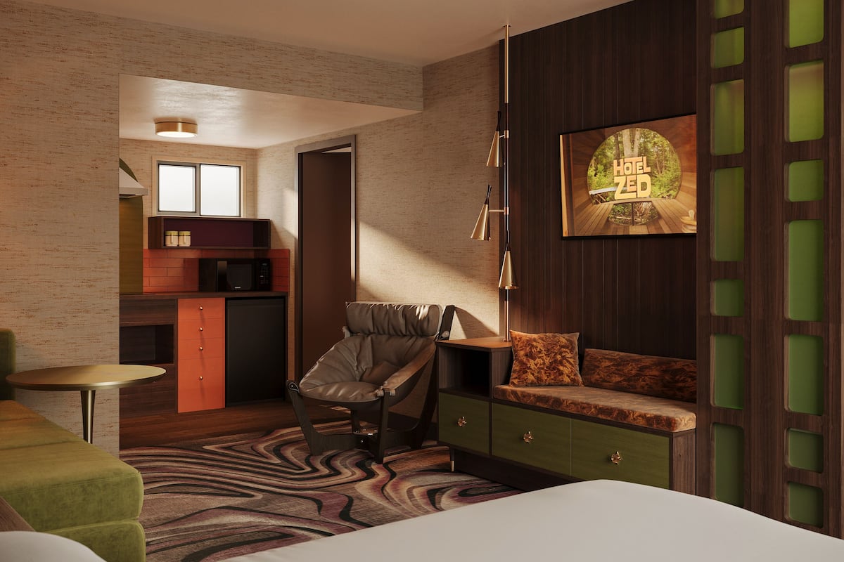 A modern hotel room with a bed, a green sofa, a chair, a desk, and a kitchen area.