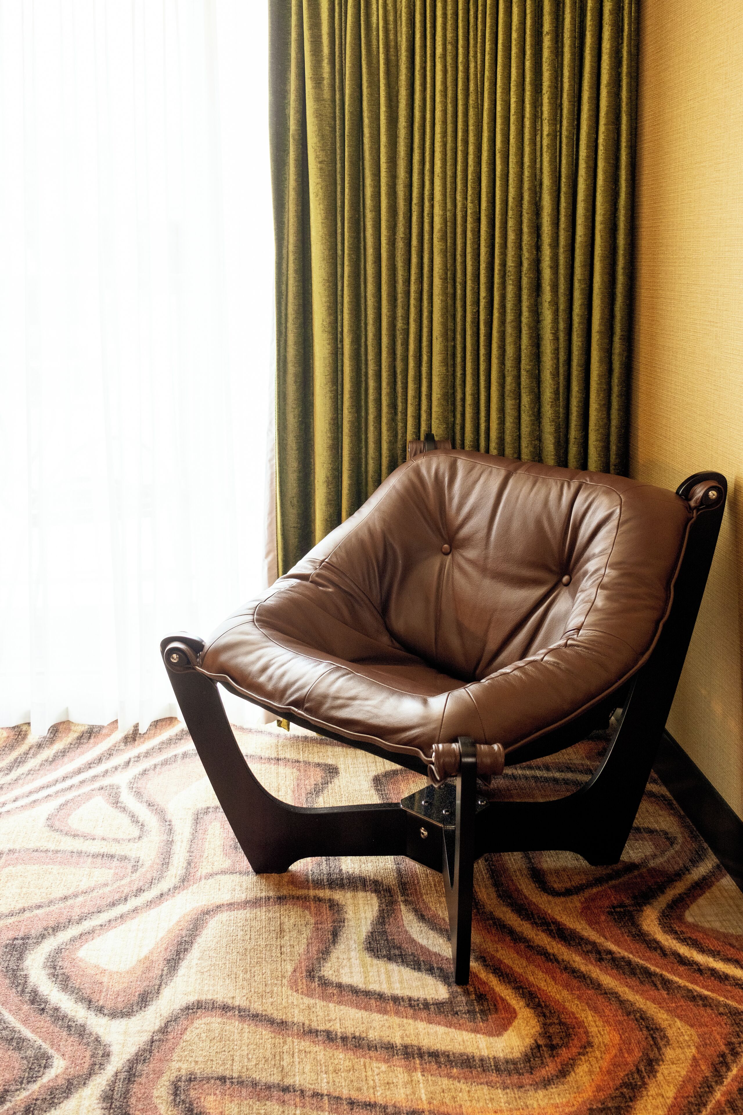 A brown leather chair with a black metal frame, positioned on a patterned carpet, next to a window with sheer curtains.