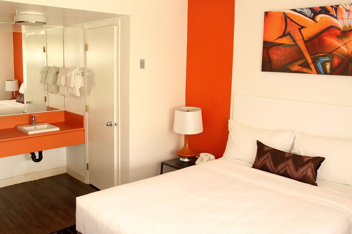 Una habitación de hotel con cama, baño con espejo, lavabo y mesita de noche con lámpara.