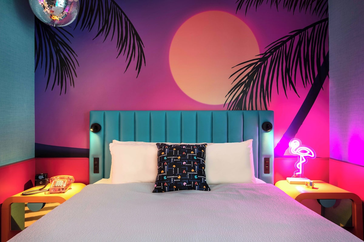 A bed with a teal headboard, a patterned pillow, and a neon flamingo sign on a bedside table.