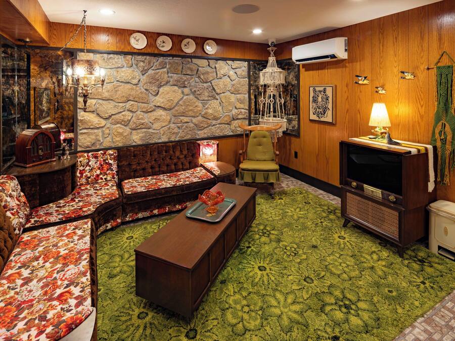 Een gezellige woonkamer met een stenen muur, een groen tapijt met patroon, een bruine bank, een groene fauteuil, een houten salontafel en een vintage radio.