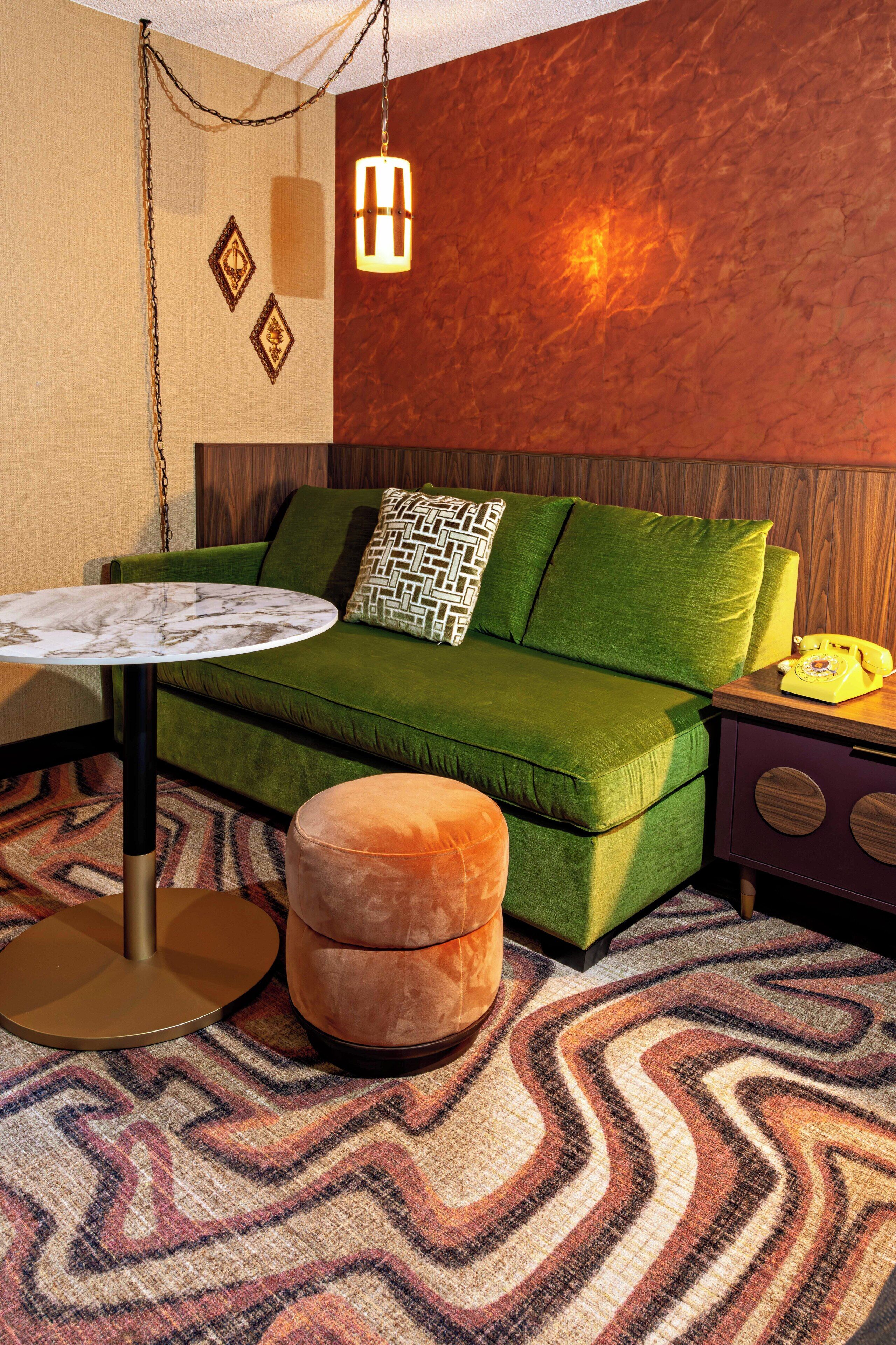 Una habitación con un sofá verde, una mesa redonda, un tapete estampado y un teléfono sobre una mesita de madera.