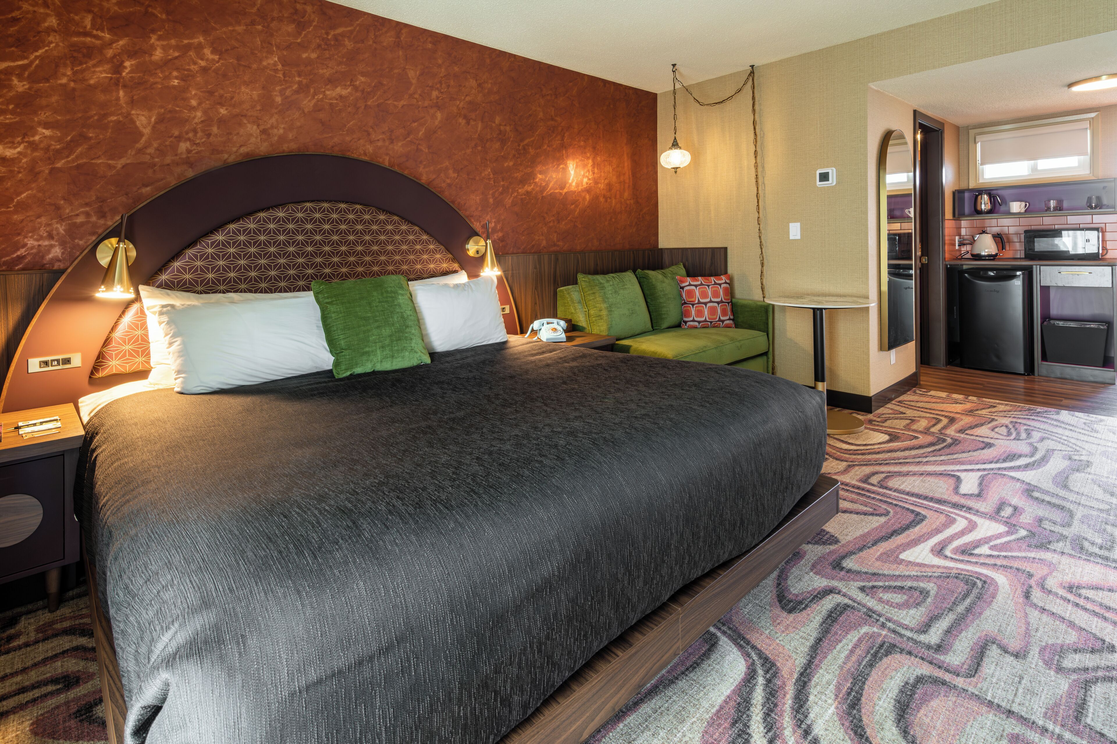 Une chambre d’hôtel moderne avec un grand lit, des oreillers verts et blancs, une table de chevet avec une lampe et un coin cuisine.