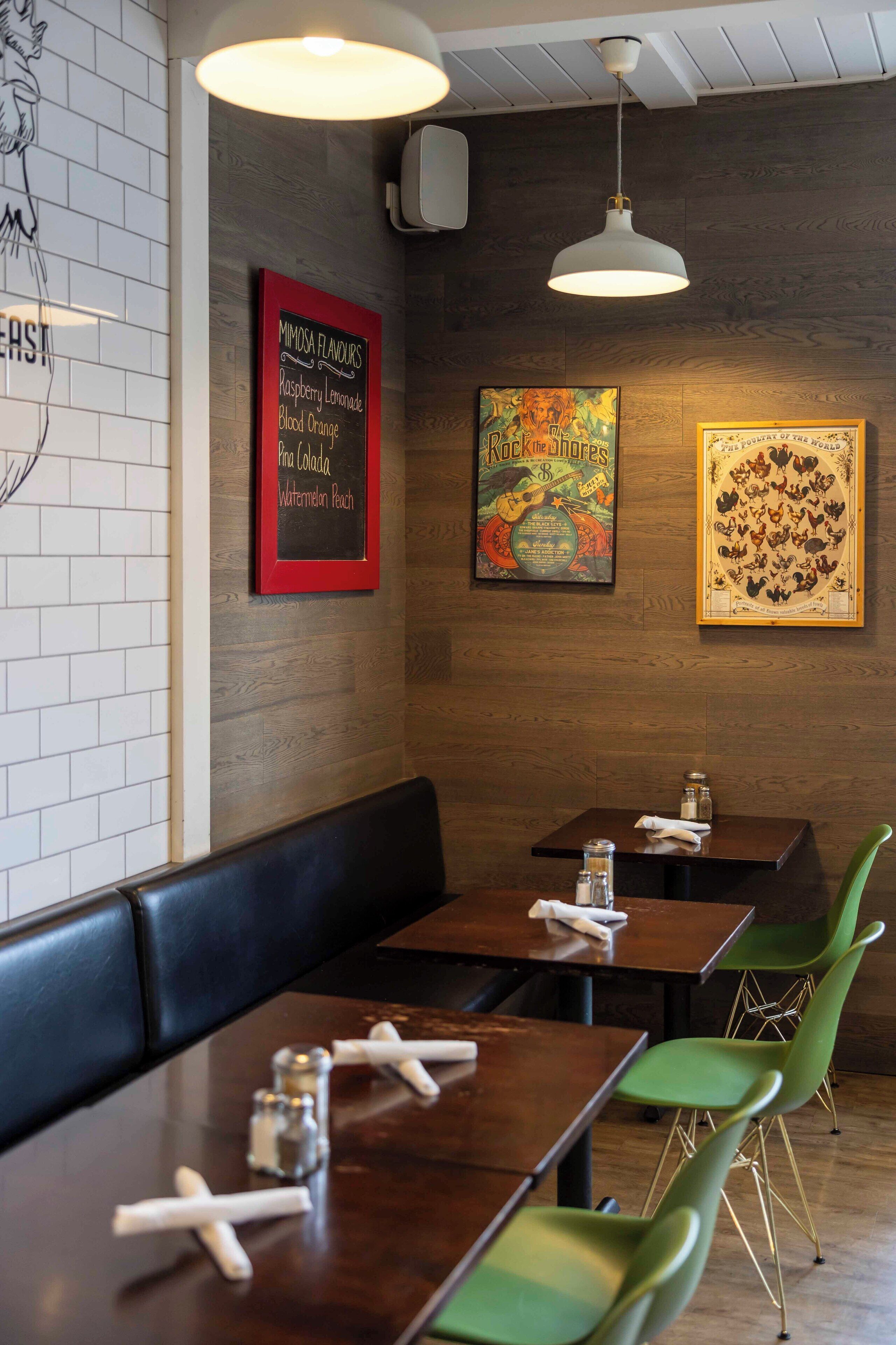 A restaurant interior with wooden tables, green chairs, and a chalkboard menu listing drinks.