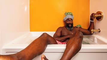 A person sitting in a bathtub with a green frog on their shoulder, a gold faucet, and a white bathtub.