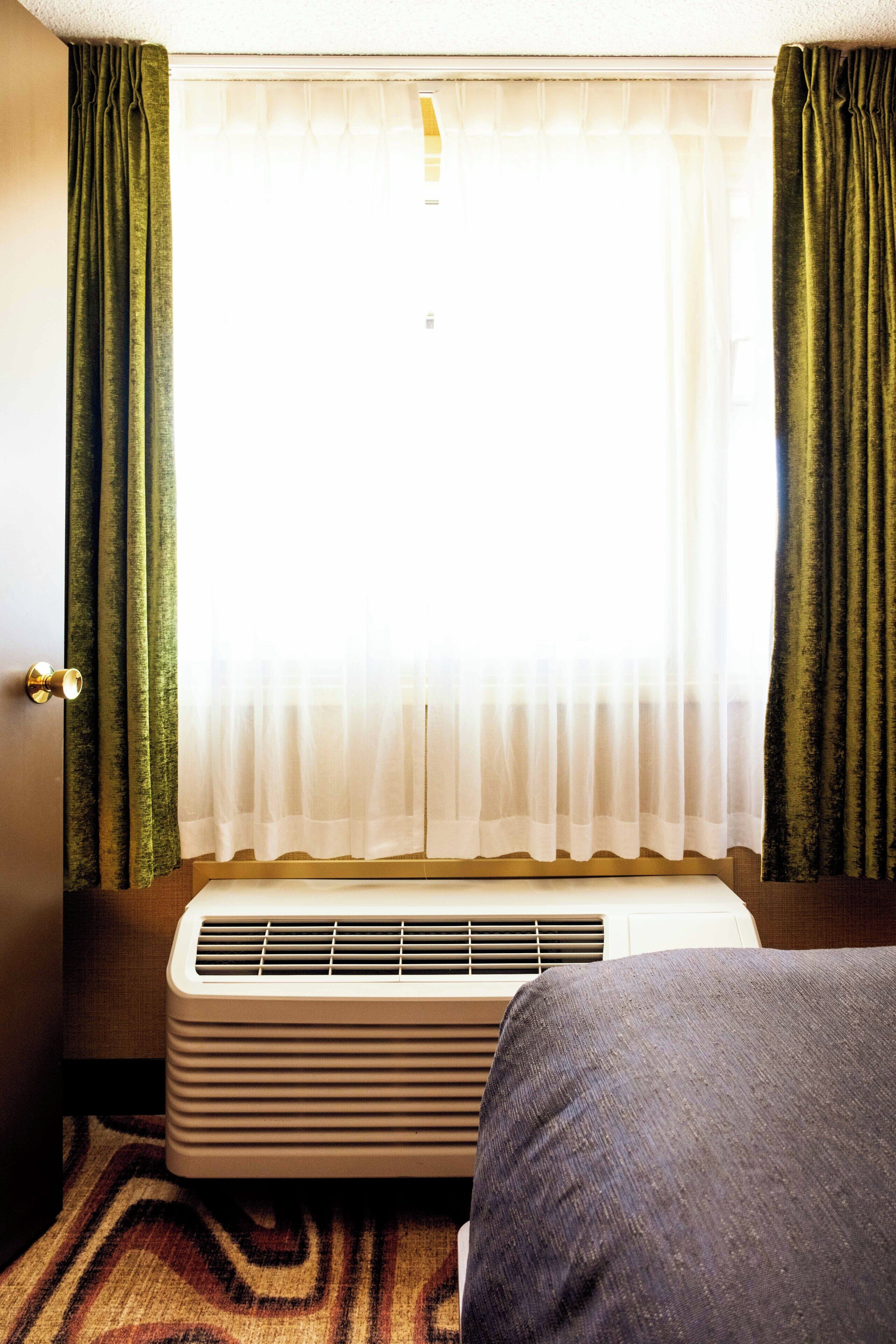 A hotel room with a bed, an air conditioning unit, a window with sheer curtains, and a door.