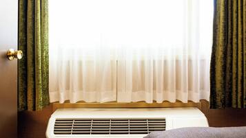 A hotel room with a bed, an air conditioning unit, a window with sheer curtains, and a door.