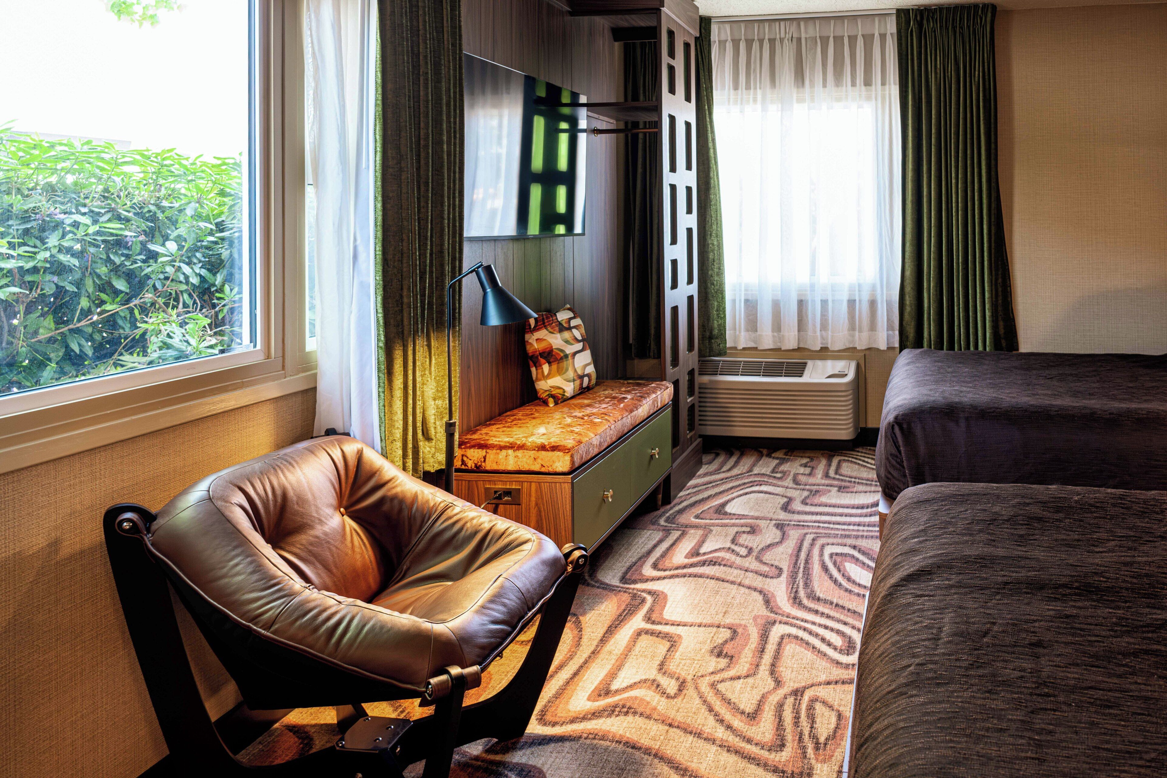 Une chambre d’hôtel avec un fauteuil en cuir, un lit, une petite table avec une lampe et une vue sur la verdure à l’extérieur.