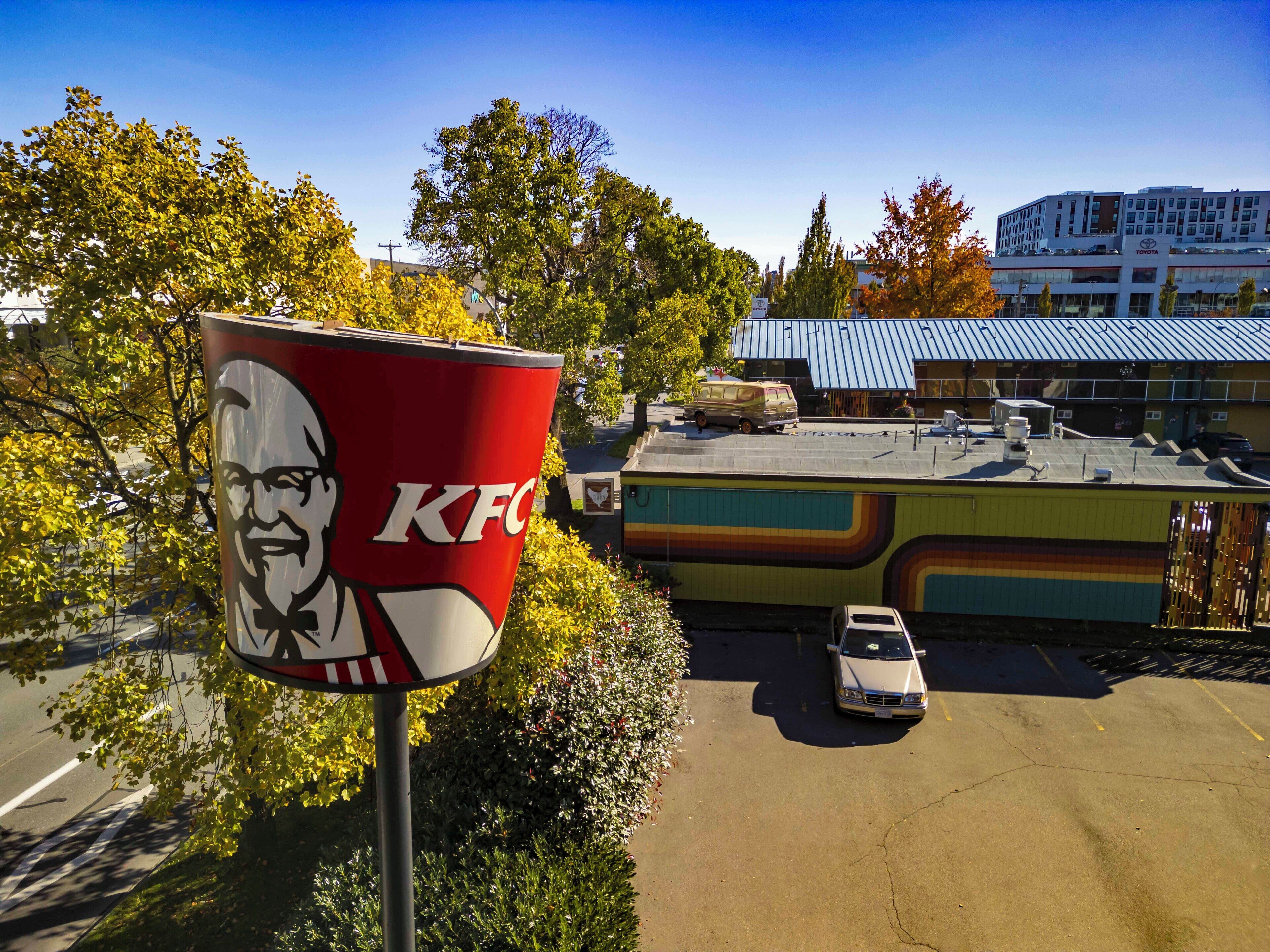 A KFC sign with a large logo, a parking lot with a white car, and a building with a blue roof in the background.