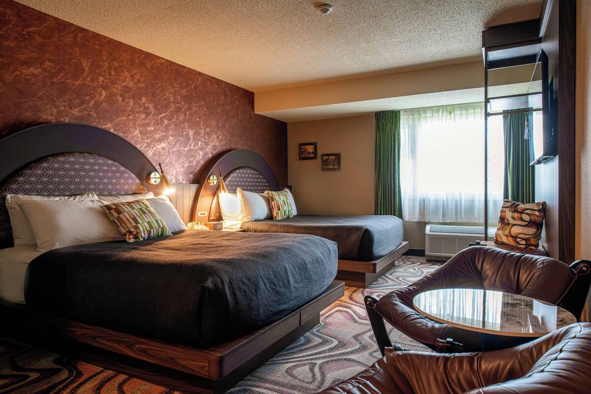 A hotel room with a large bed, a smaller bed, a leather armchair, a glass table, and a large window with curtains.
