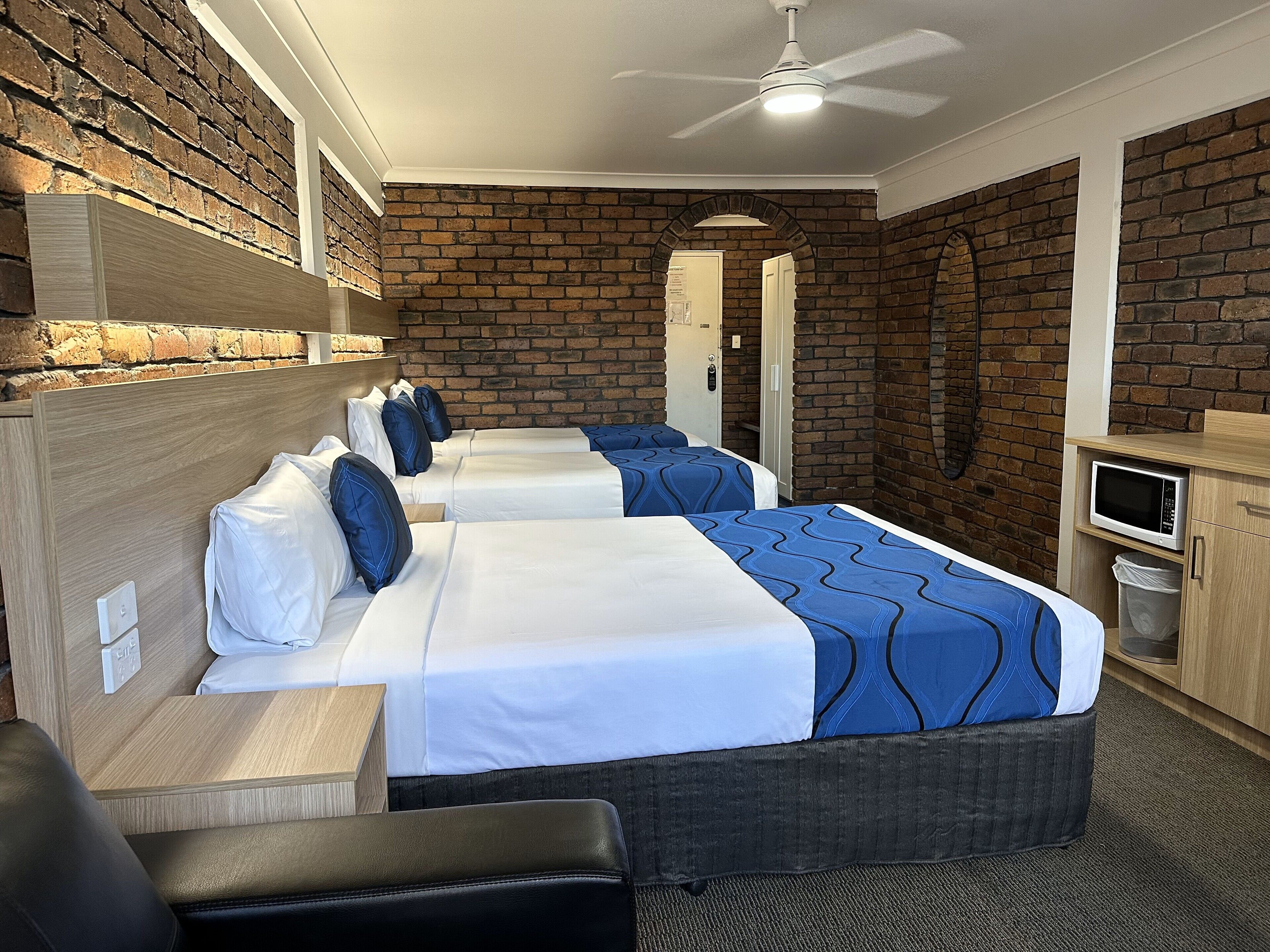 A hotel room with three beds, a ceiling fan, a microwave, and a brick wall.